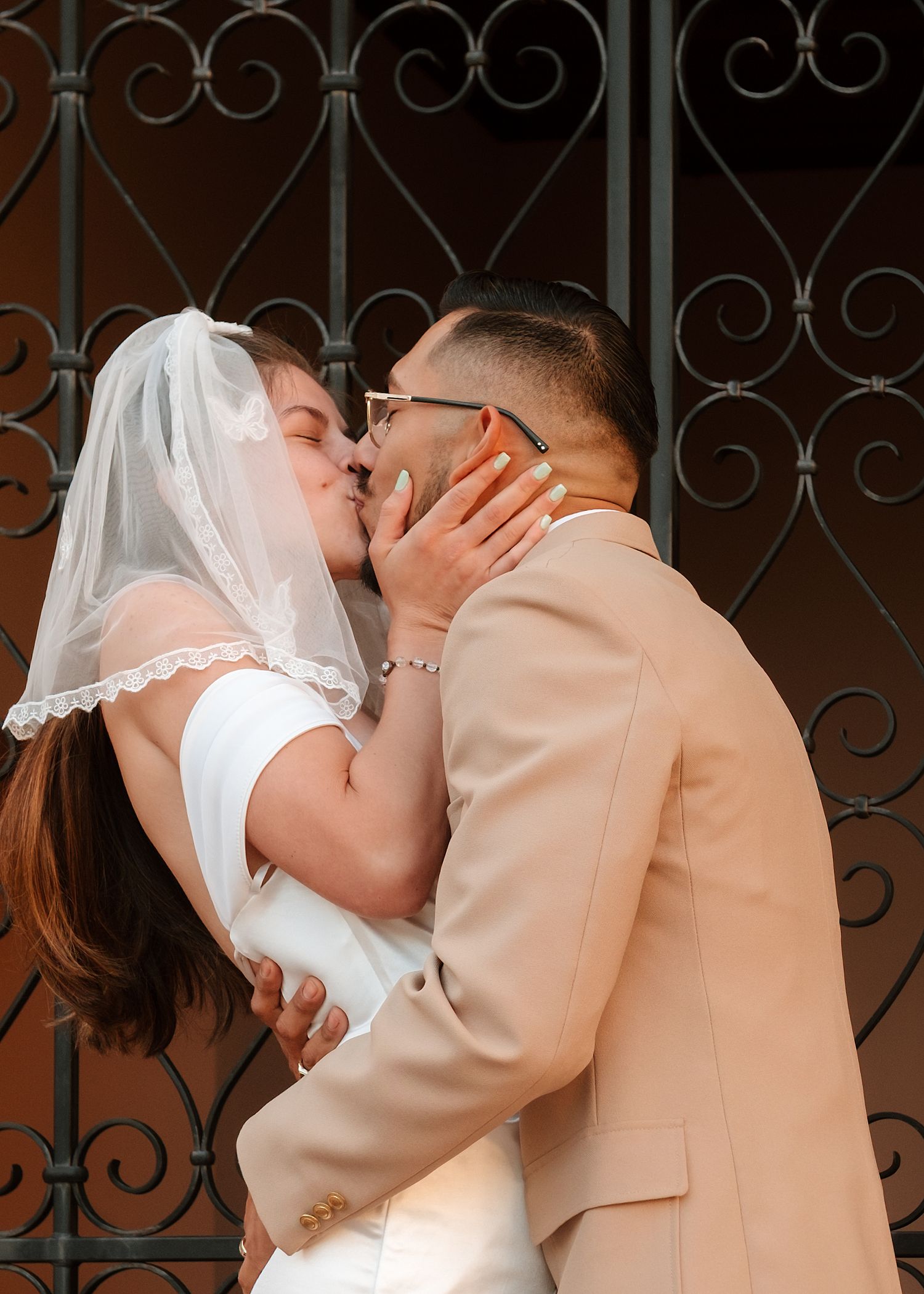 A bride and groom share a romantic kiss in front of ornate iron gates, bride wearing veil and white dress.