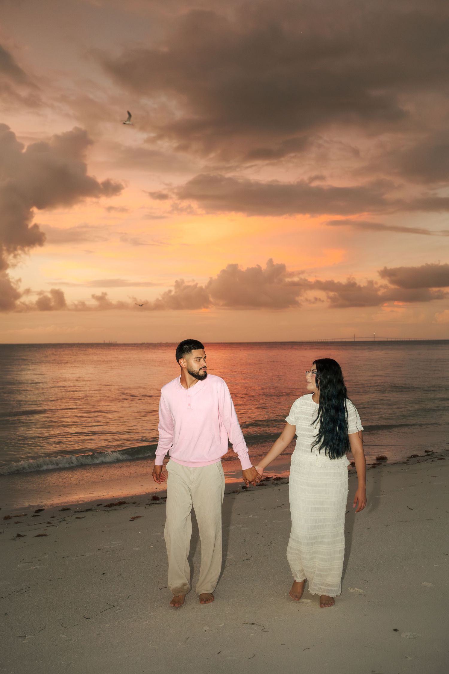 Couple holding hands on a beach at sunset, man in pink shirt and woman in white dress, dramatic cloudy sky.