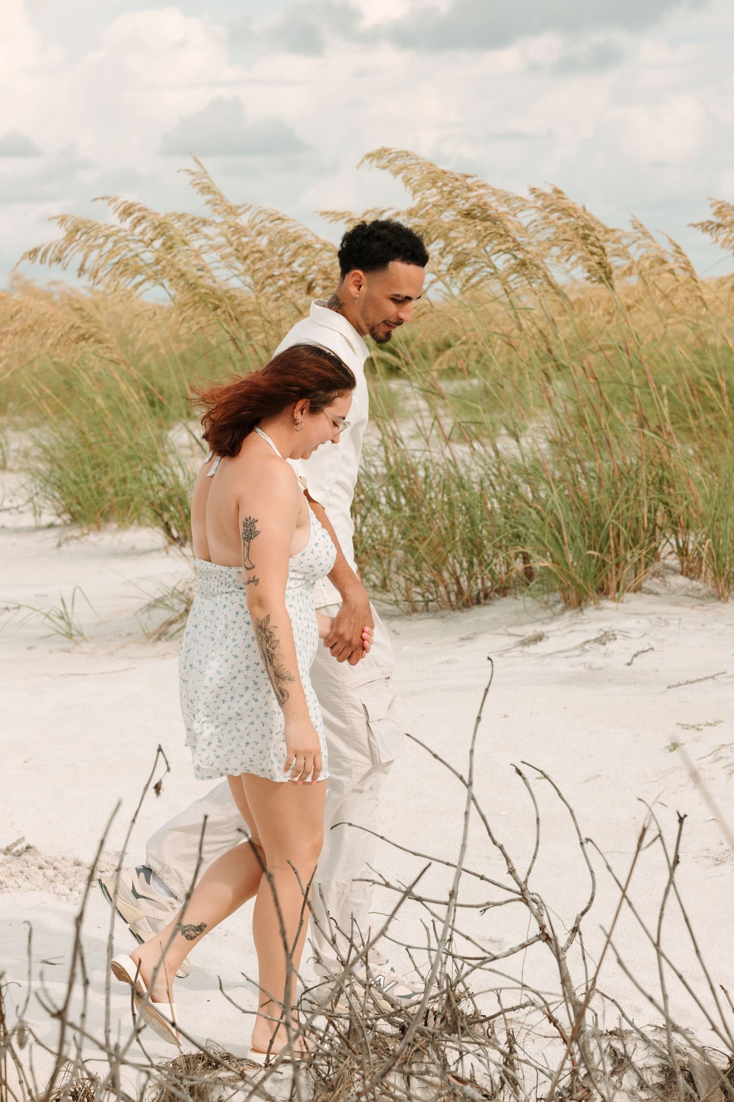 Couple holding hands walking on white sand dunes with sea oats, beach engagement photo session.