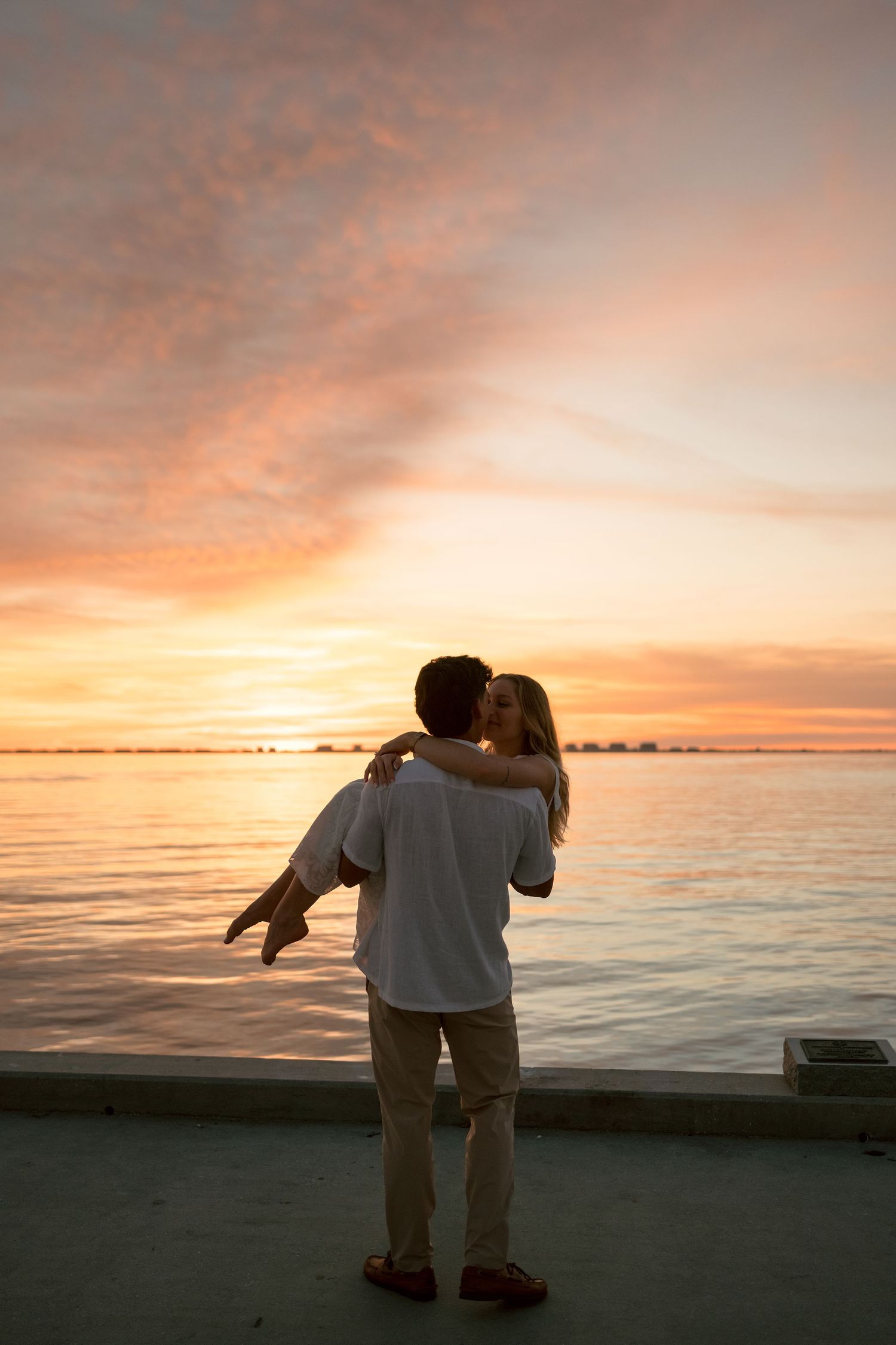 Couple sharing a romantic kiss at sunset by the waterfront, with a stunning orange and pink sky reflecting on calm waters.