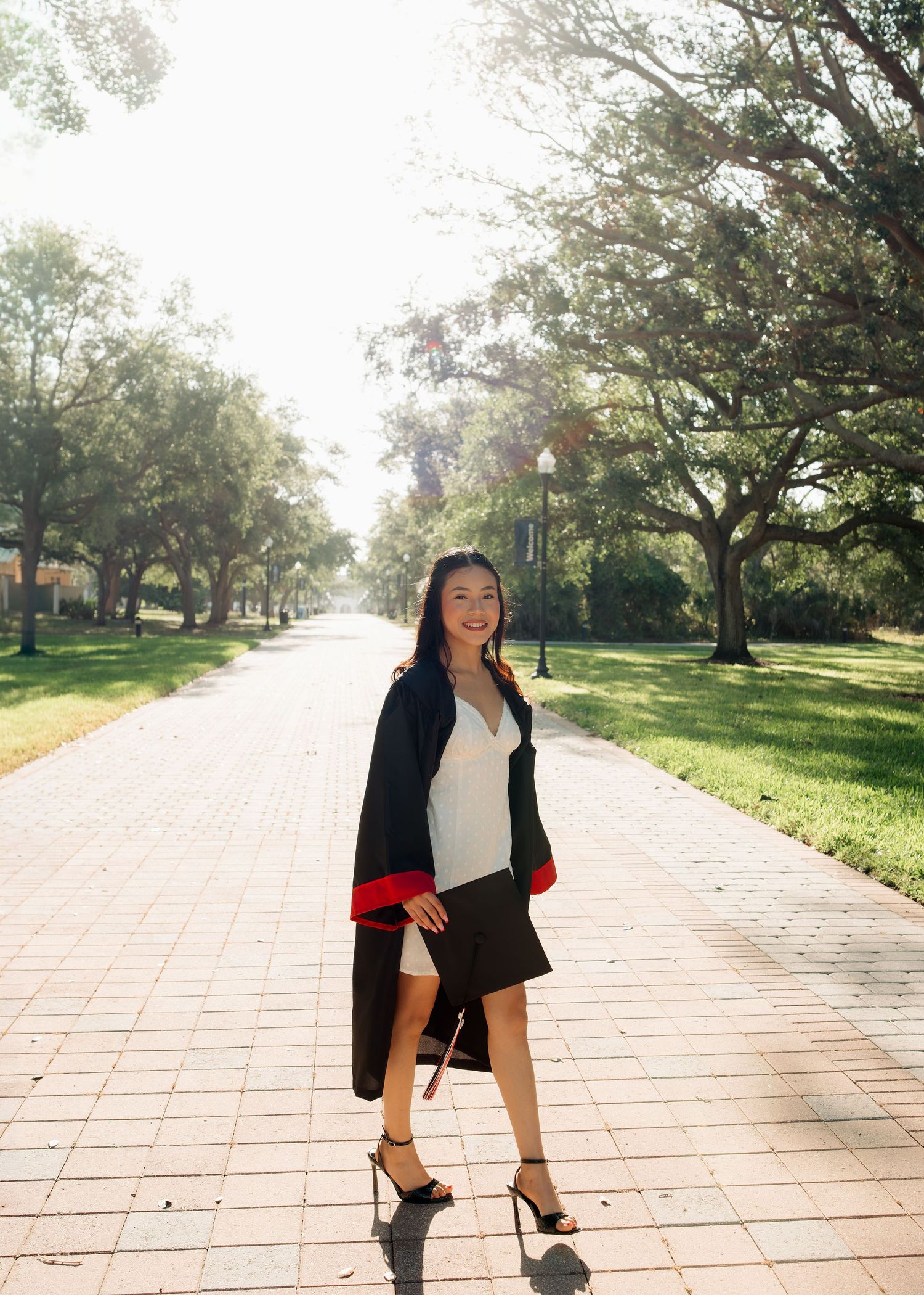 A smiling young woman in a white dress and black graduation gown walks along a sunny tree-lined brick pathway.