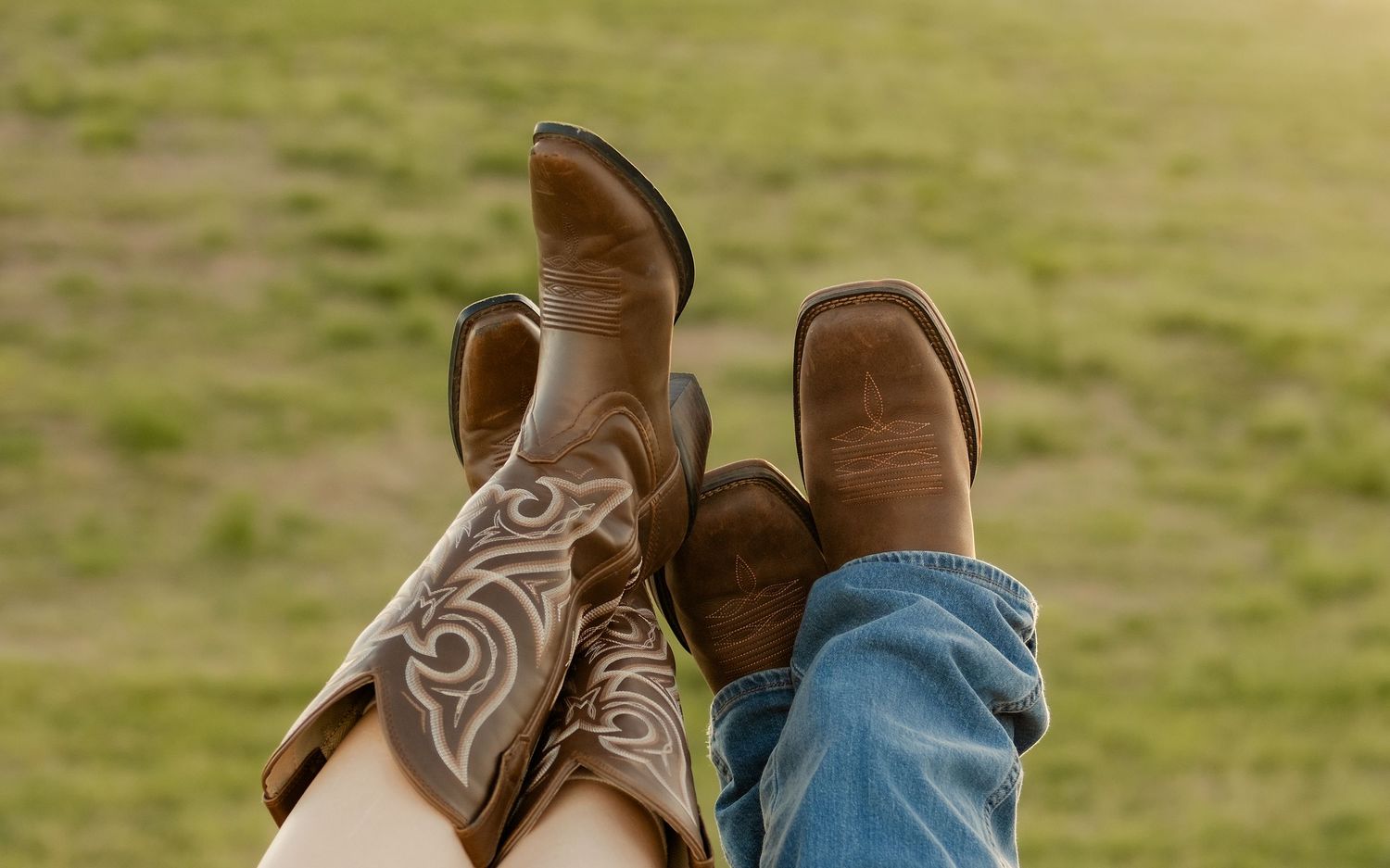 Two pairs of cowboy boots with decorative stitching raised up against a blurred green grass background.