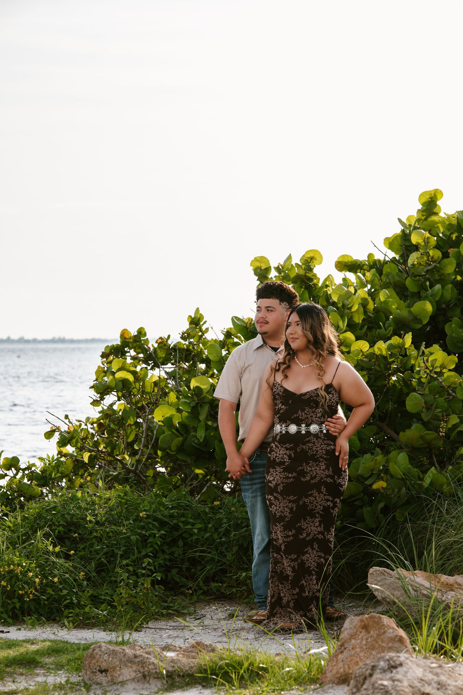 Couple standing together near lush green tropical foliage by the ocean, woman in floral dress, man in casual shirt.