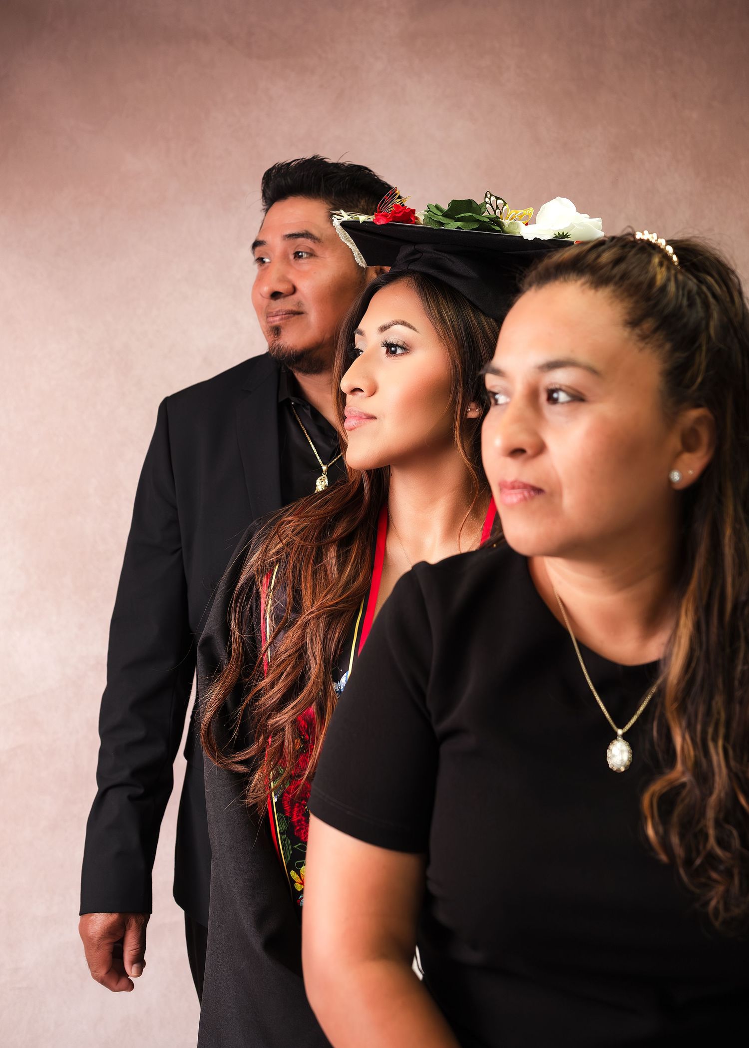 Three people in black attire pose for a studio portrait, two women wearing floral crowns and a man in a suit jacket.