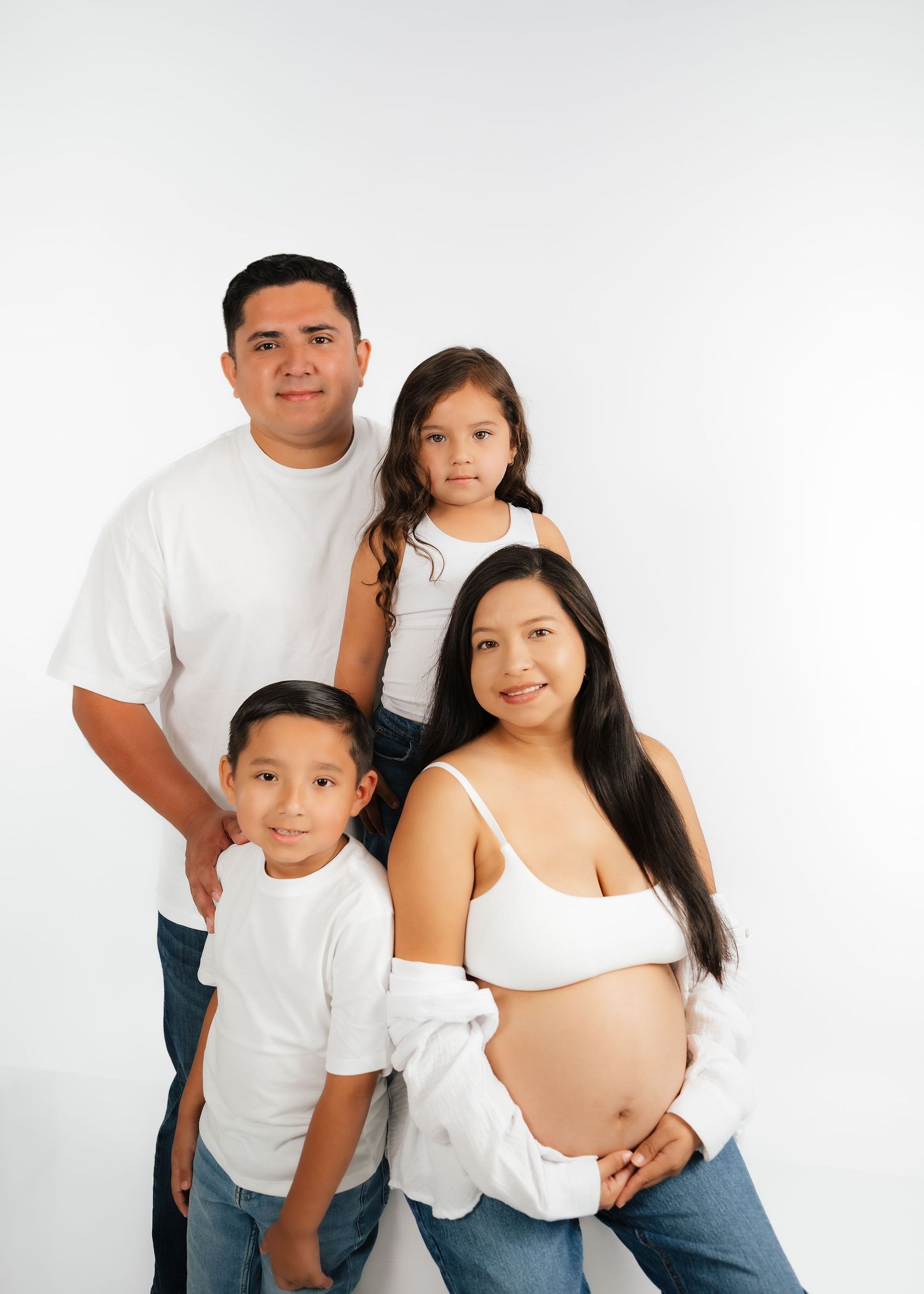 Family maternity photo with pregnant mother, father, and two children in white tops and jeans on white background.