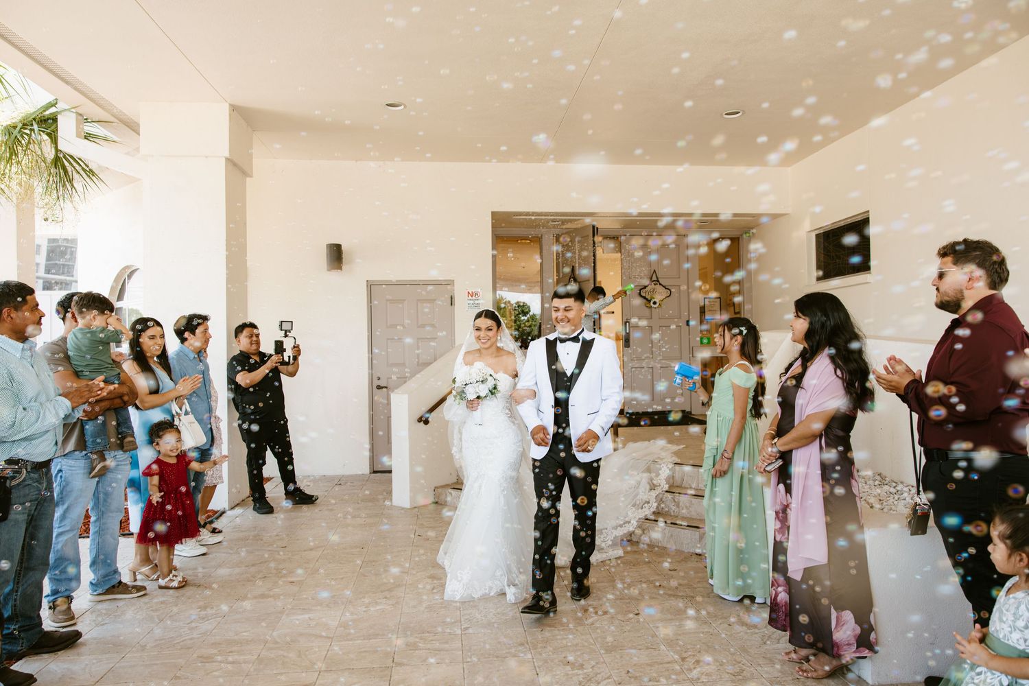 Bride and groom walk through bubble send-off surrounded by smiling guests at outdoor wedding venue.