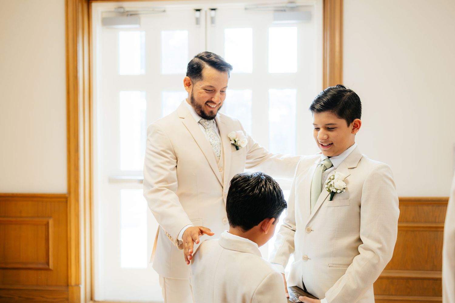 Three men in white suits share a joyful moment together before a wedding ceremony indoors.