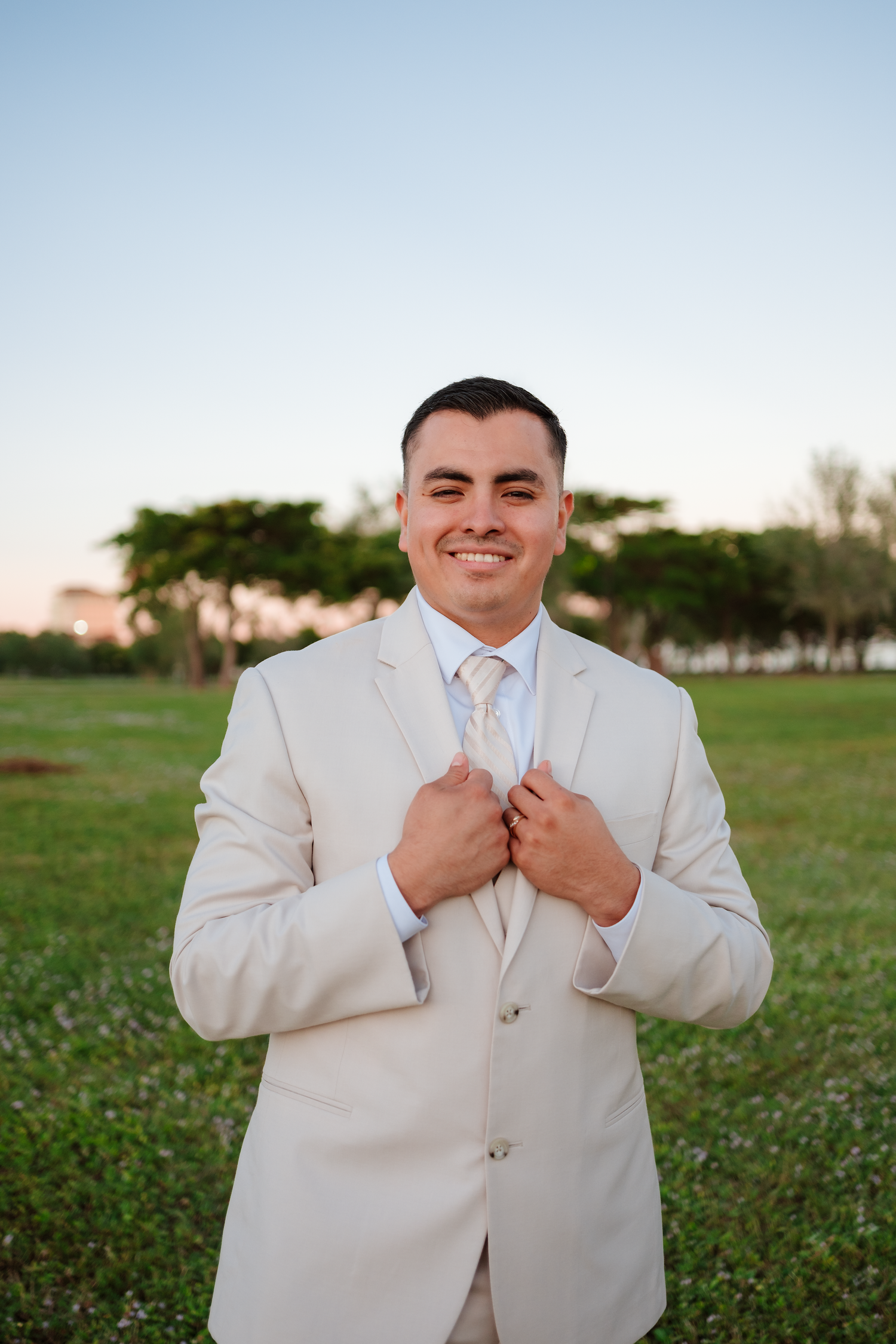Smiling groom in tan suit adjusting lapels outdoors on green lawn at dusk with trees in background.
