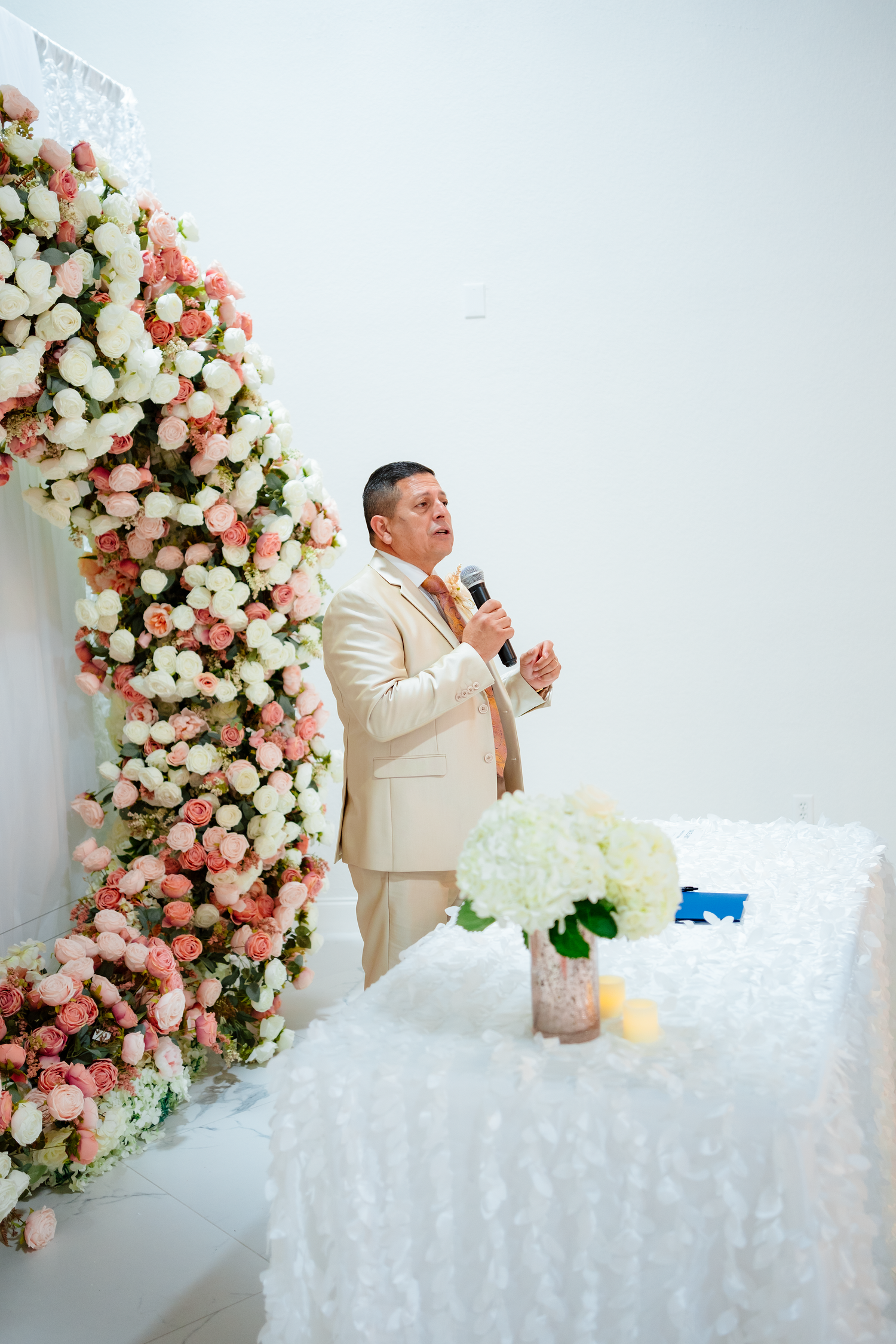 Officiant in cream suit speaking into microphone at elegant wedding ceremony with pink and white floral arch.