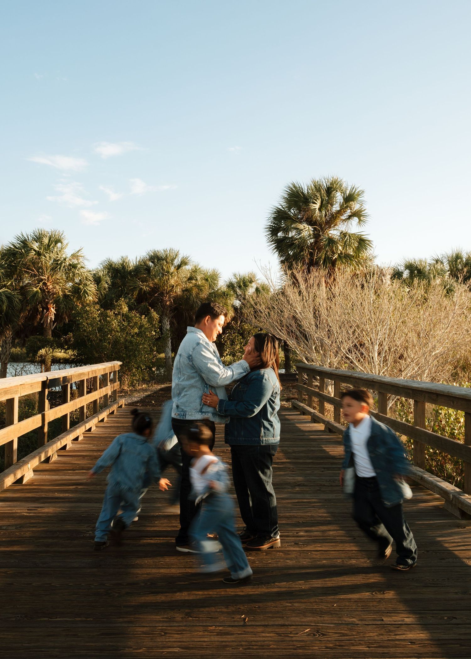 Family with children running on a wooden boardwalk surrounded by palm trees at golden hour.