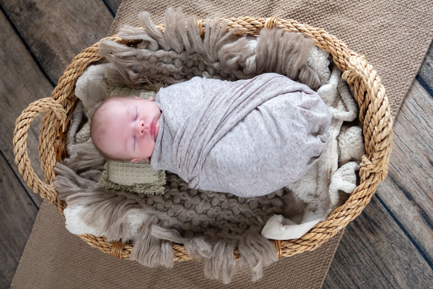 Sleeping newborn wrapped in gray swaddle rests in woven basket with fur lining.