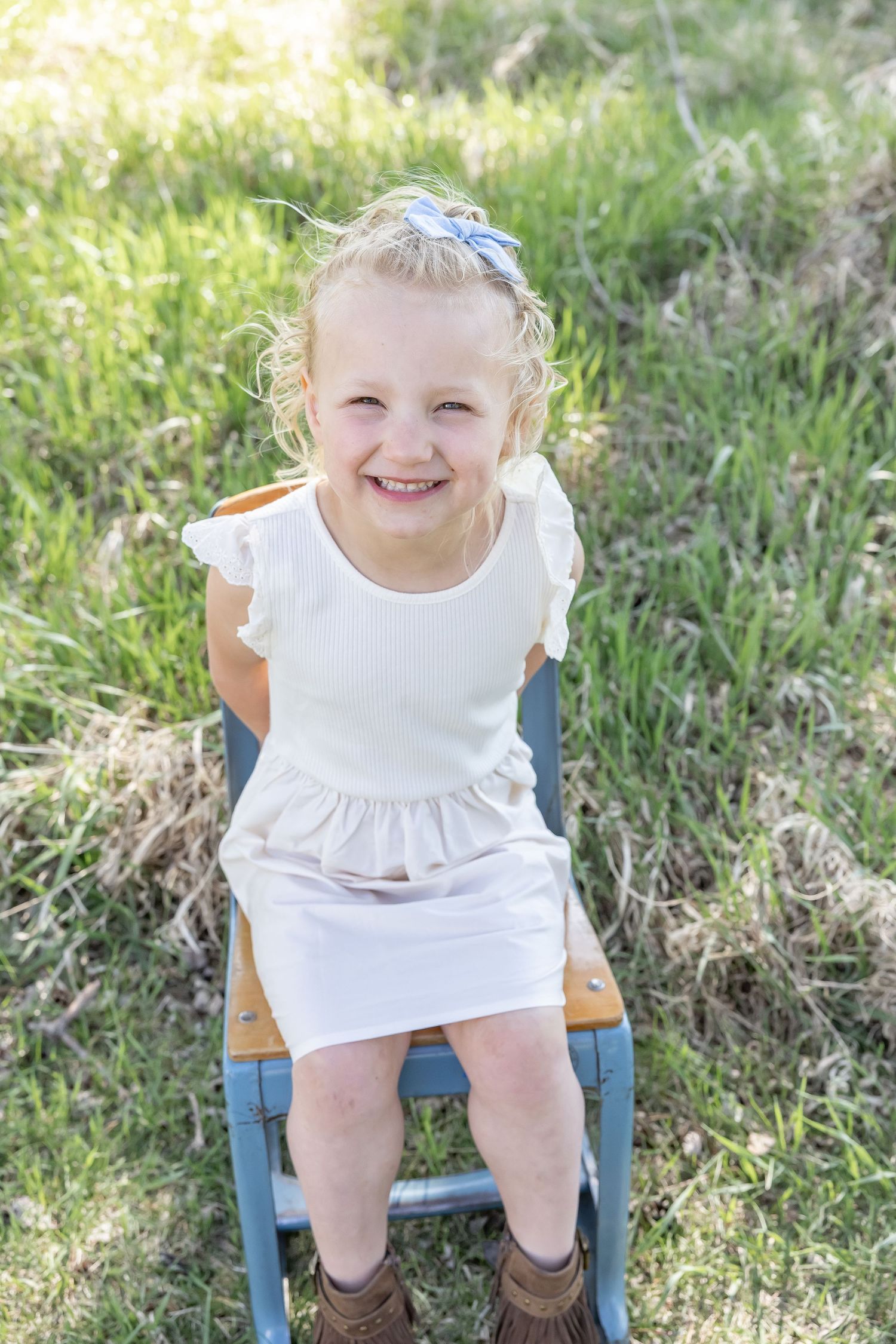 Child in white ruffled dress sits on wooden chair in grassy field.
