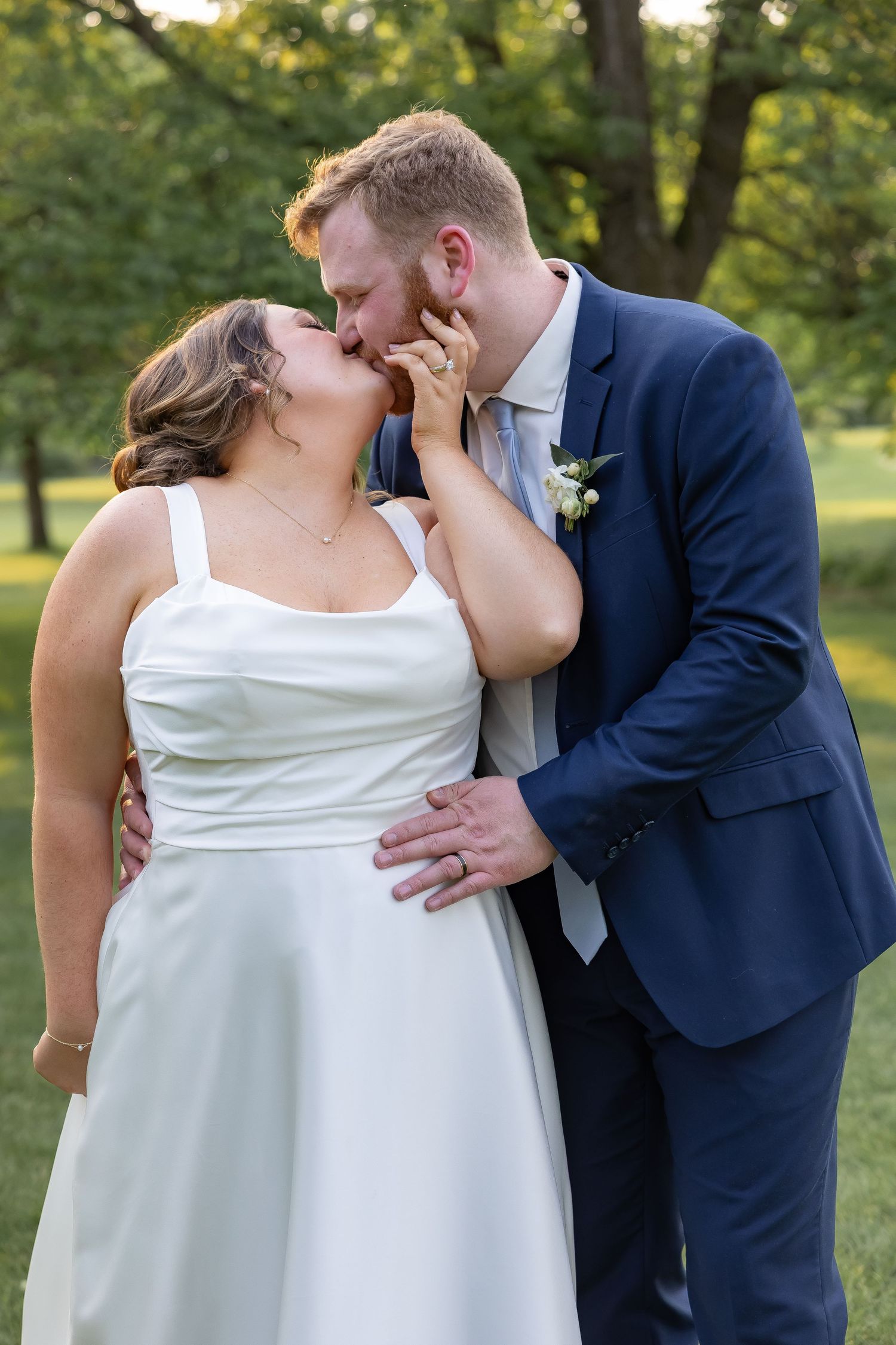 A couple in wedding attire share an intimate moment outdoors with trees and greenery in the background.