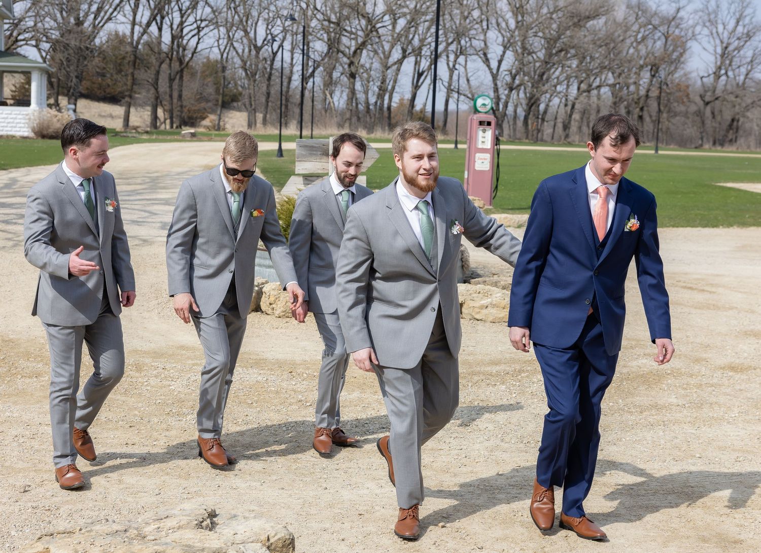 Groomsmen in grey and blue suits walking in a line on a gravel path.
