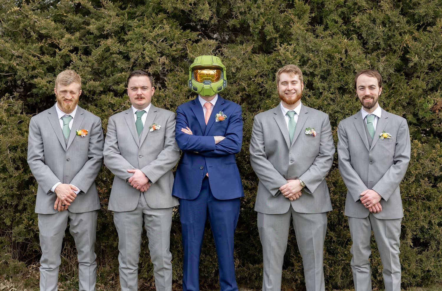 Wedding party members in grey and navy suits lined up against greenery for formal portraits.