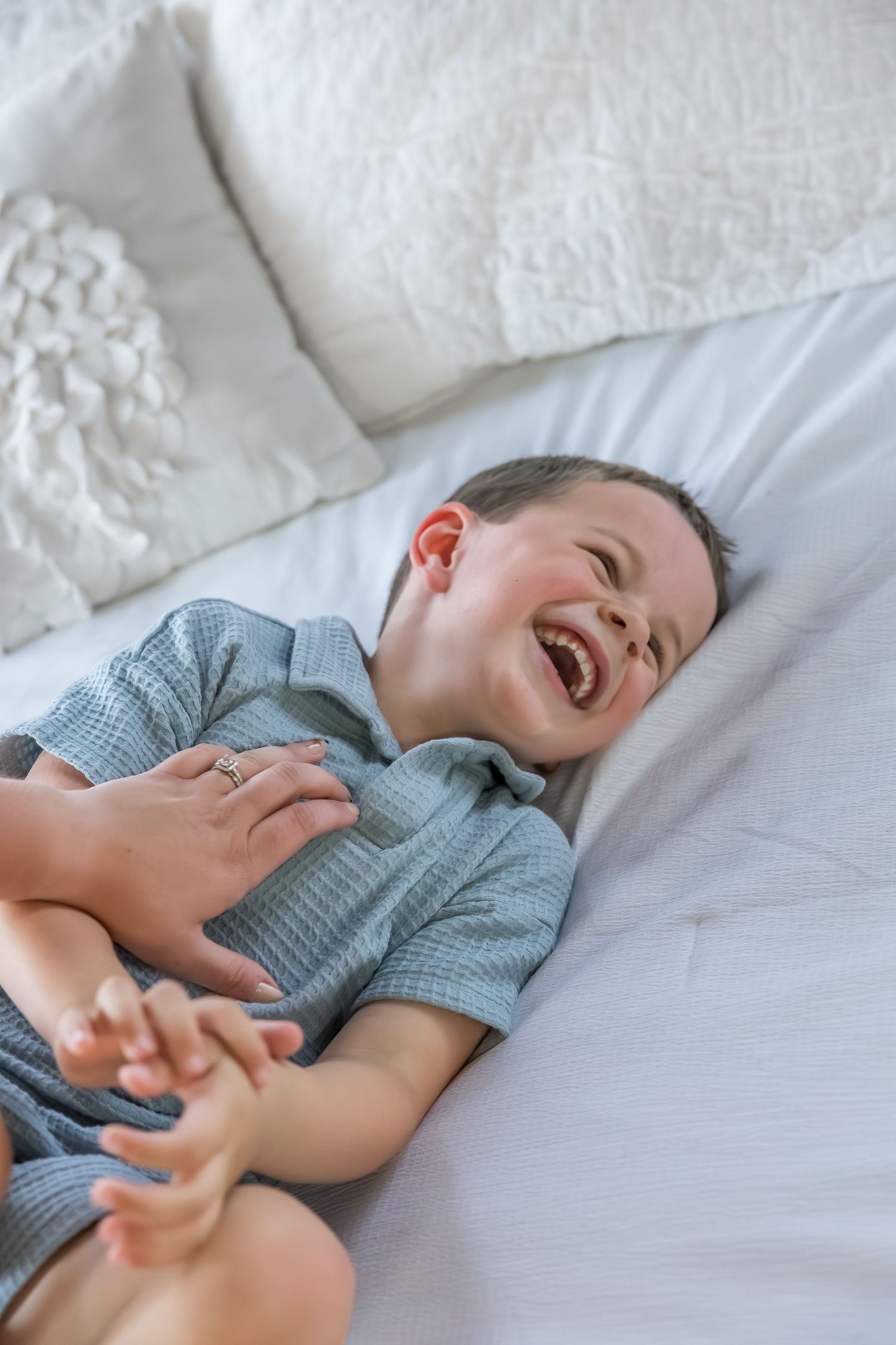 A child in blue clothing lies on white bedding while laughing heartily.
