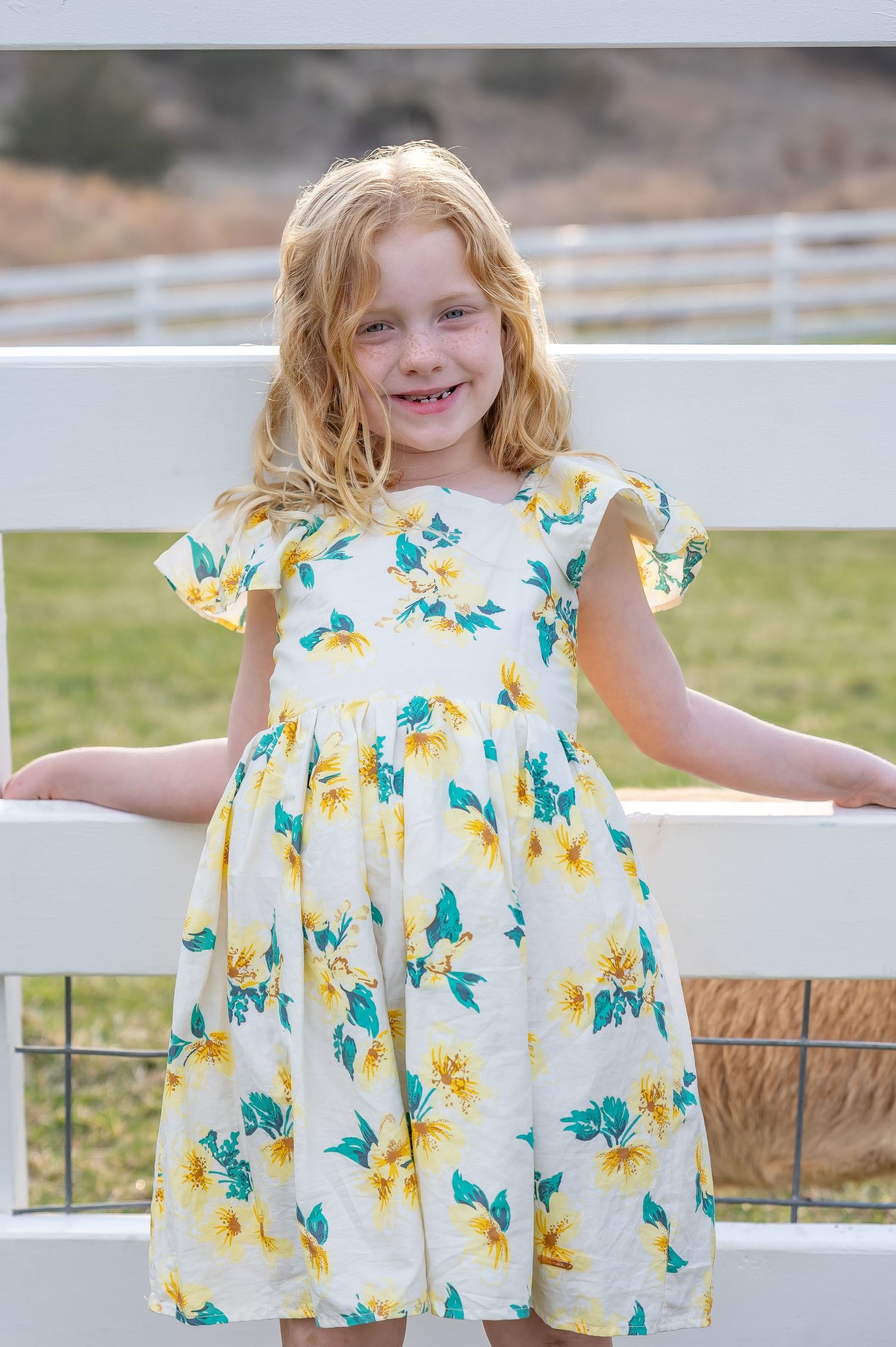 A young child wearing a white and yellow floral dress stands outdoors near a white fence.
