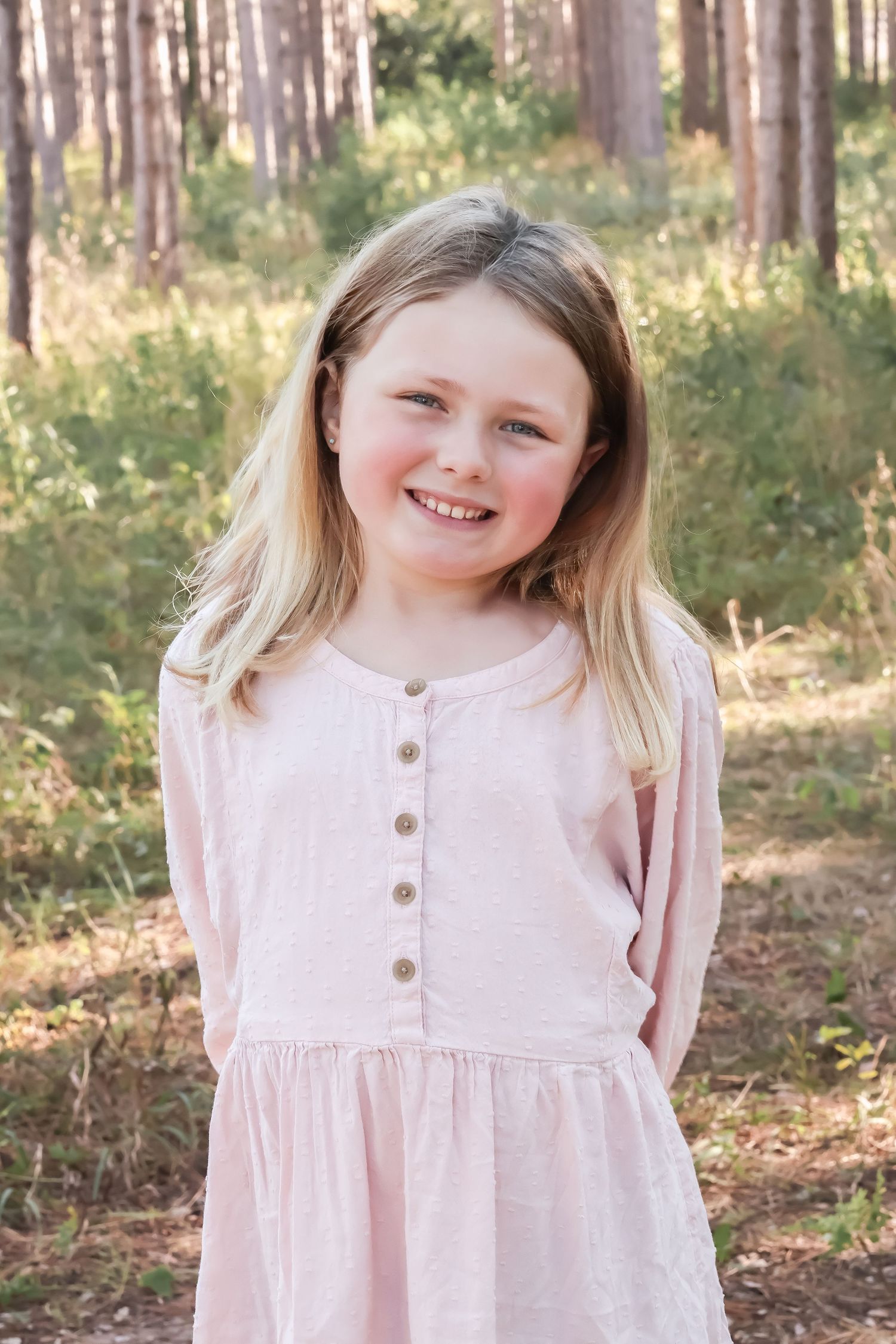 Child in pink button-front dress poses outdoors among pine trees.