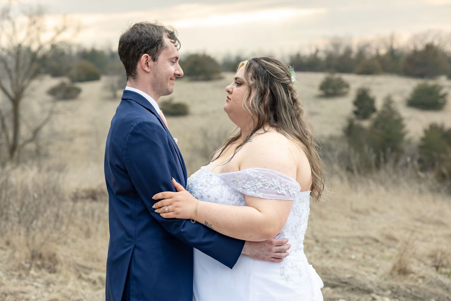 Wedding couple embracing in an outdoor field setting wearing navy suit and white gown.