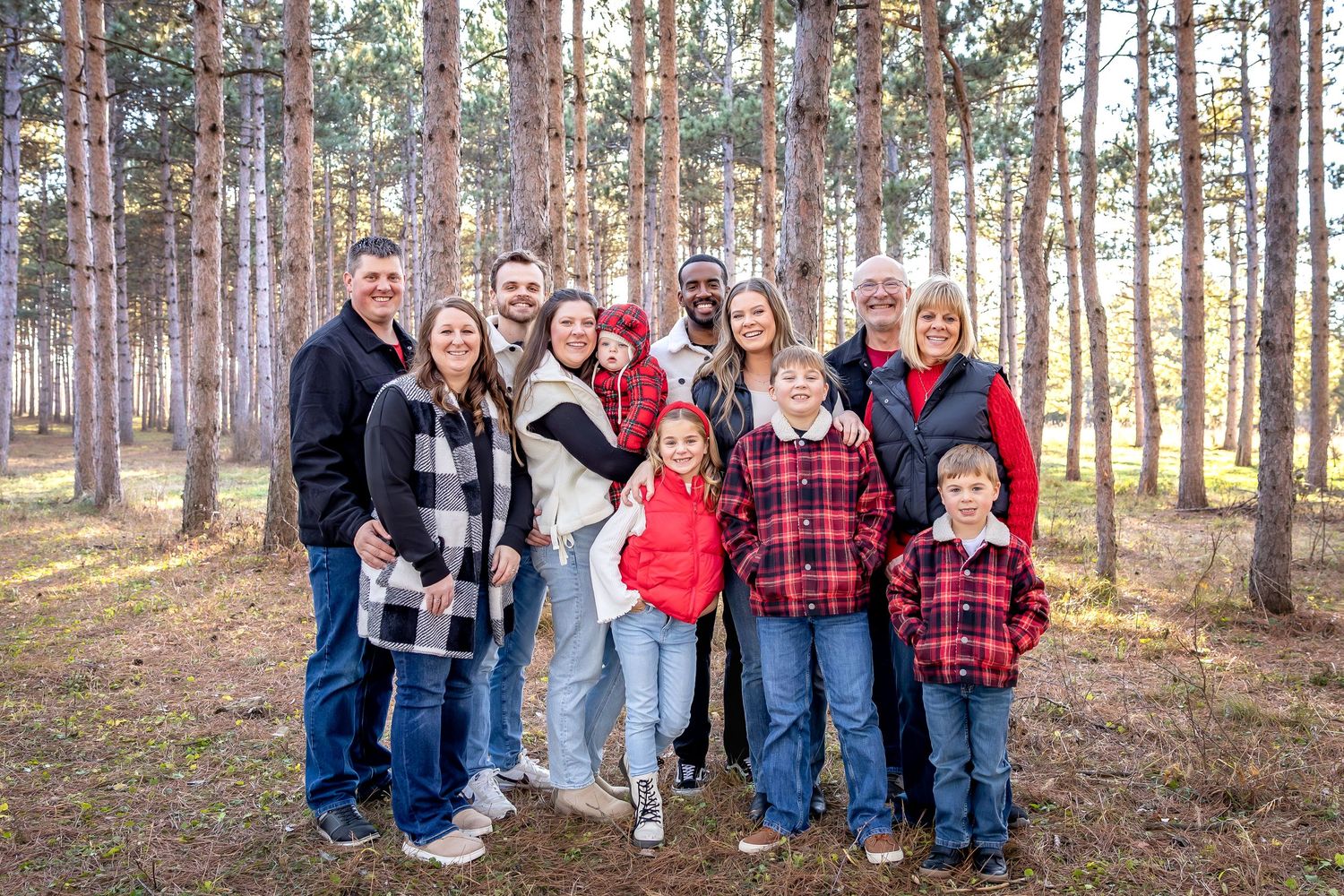 A group of people in casual winter attire pose together in a pine forest setting during golden hour.