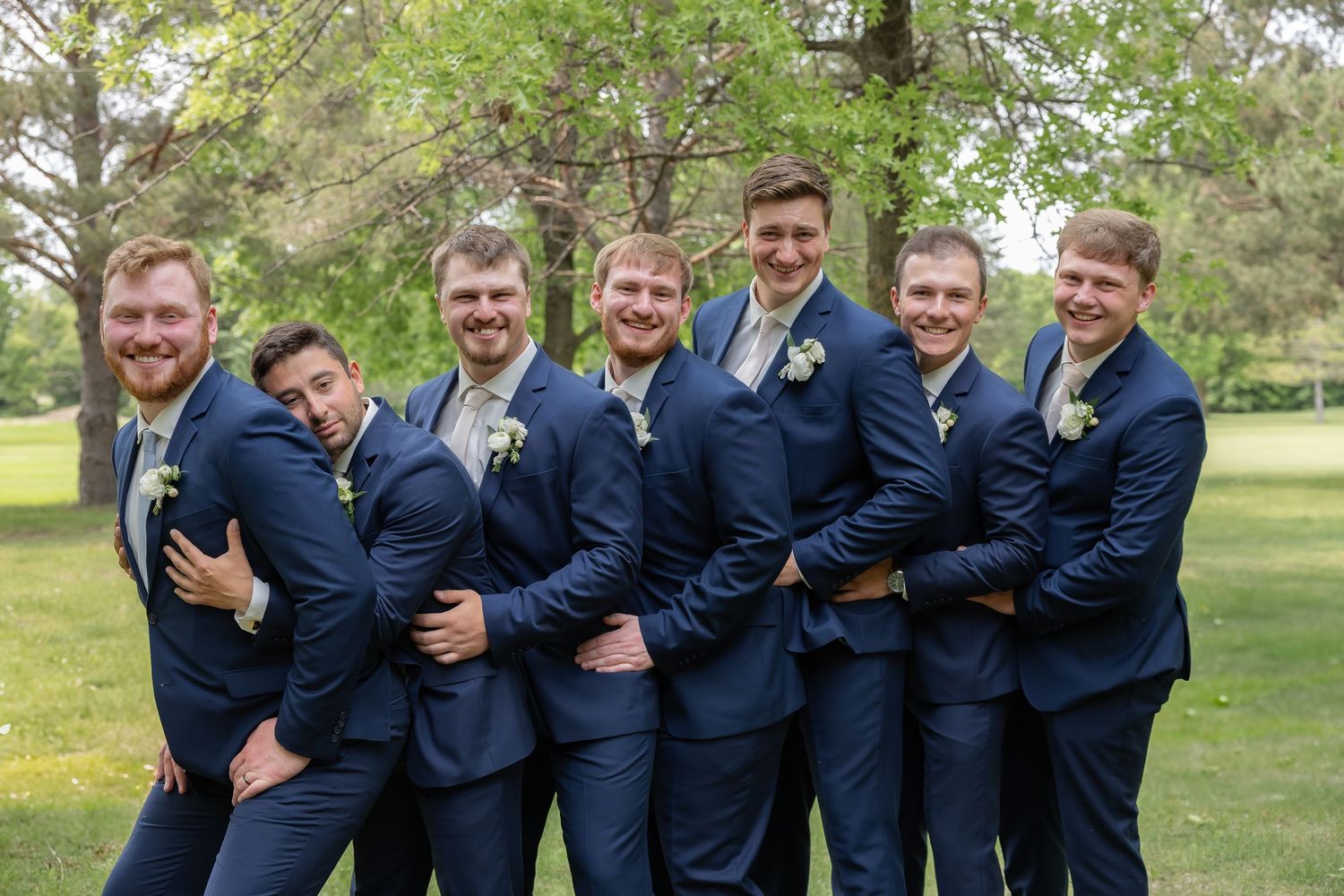 A group in matching navy suits pose together outdoors in a park setting for a formal event photo.
