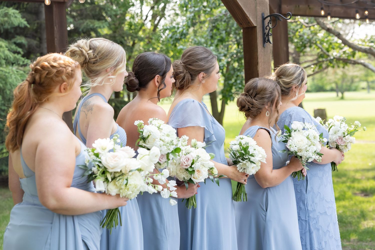 Bridesmaids in light blue dresses stand together holding white bouquets outdoors.
