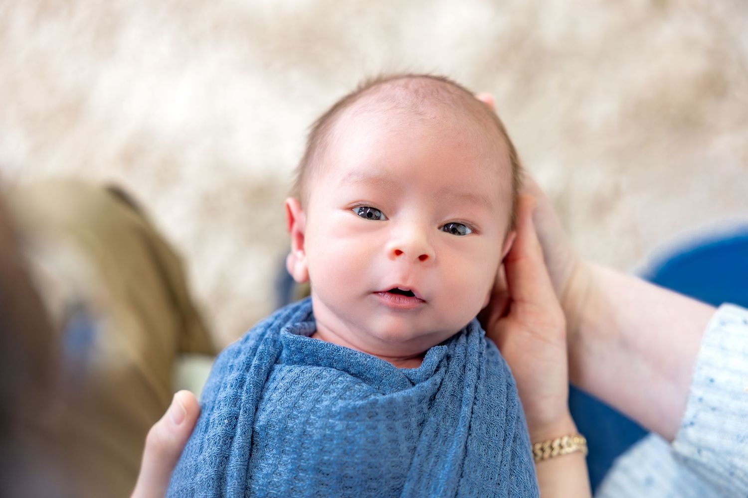 A baby wearing a blue knit sweater smiles while lying on a white blanket.