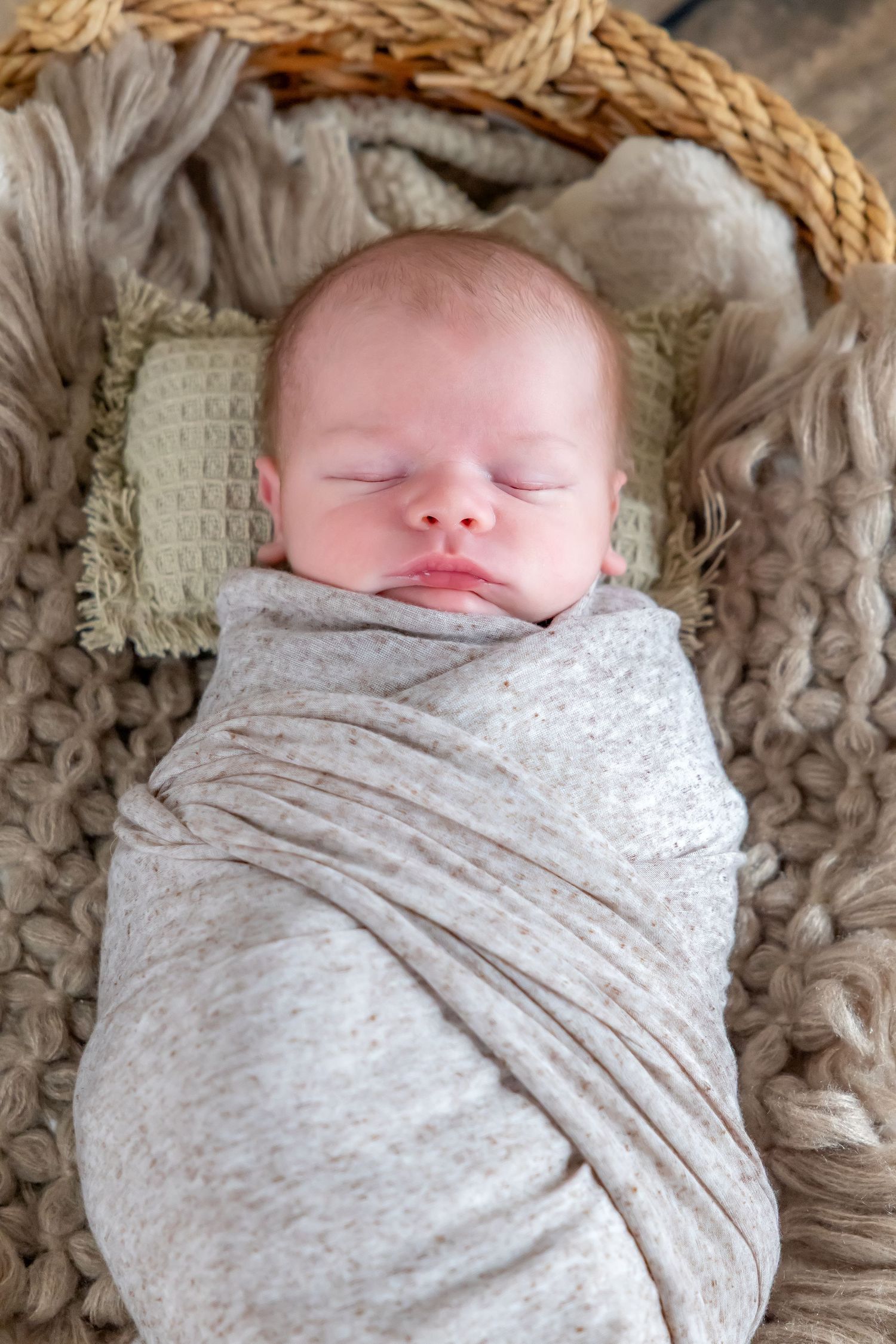 Peaceful newborn swaddled in gray wrap sleeps in woven basket on textured blanket.