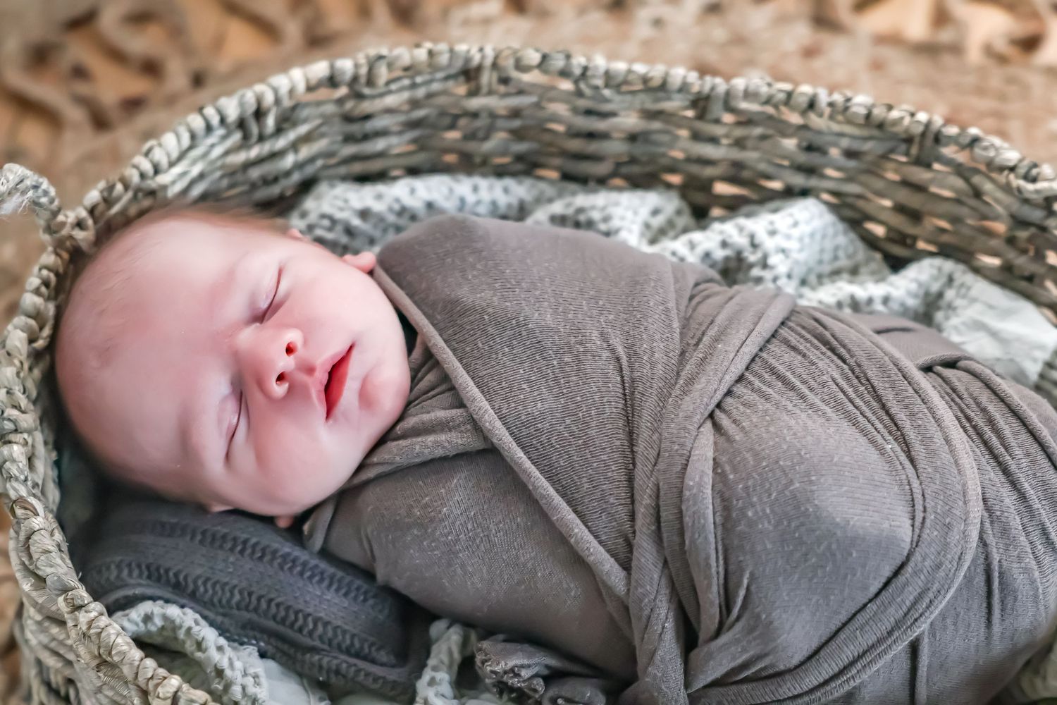Sleeping baby wrapped in gray swaddle resting in woven basket.