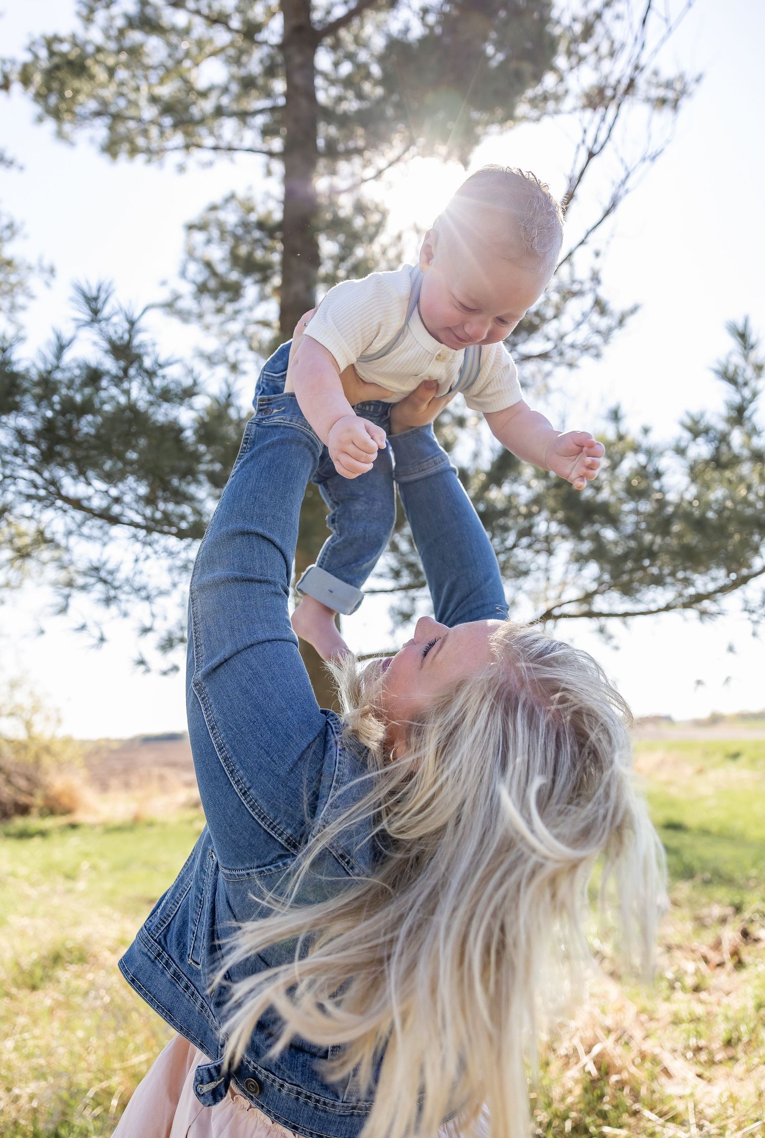 Parent lifts joyful toddler in air during playful outdoor photoshoot with sun flare.