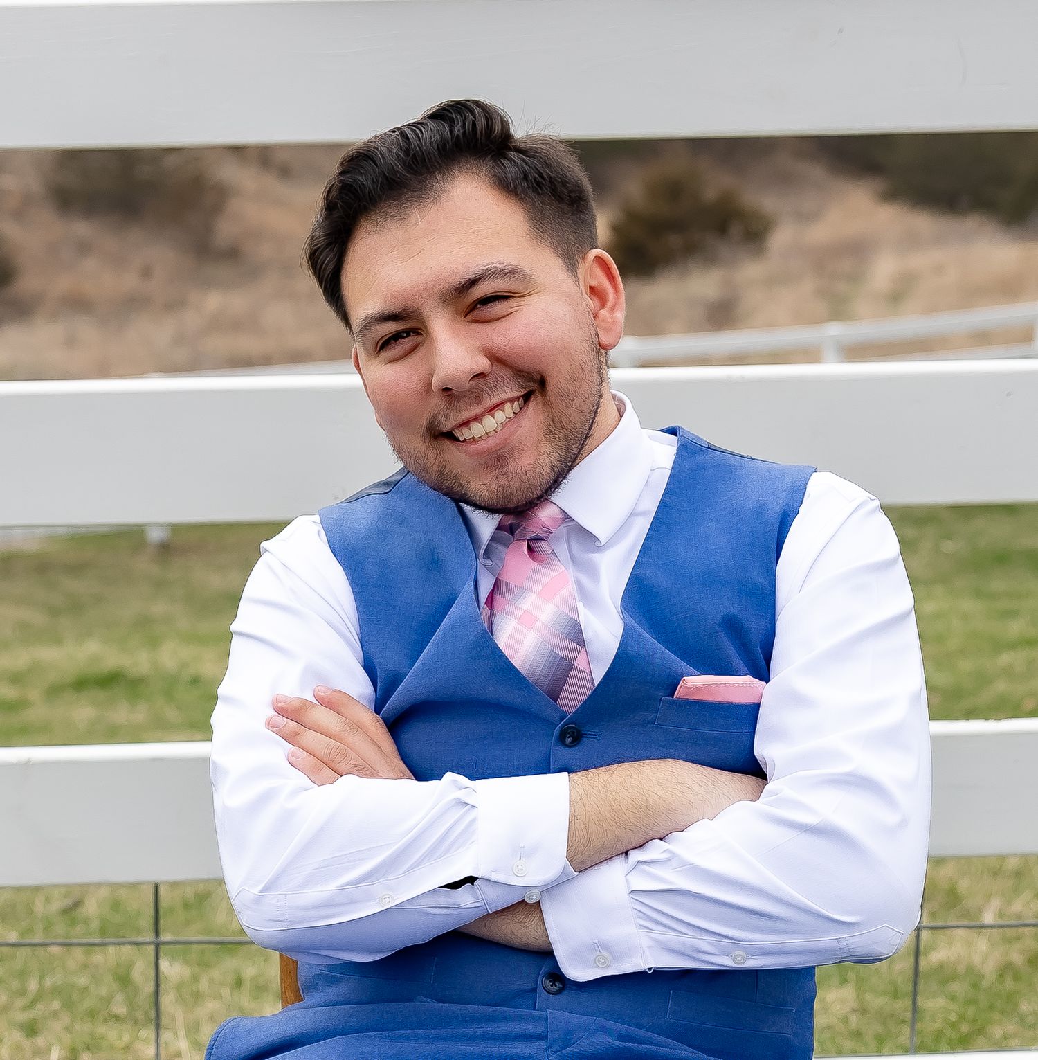 Person in blue vest and pink tie smiling confidently against rural background with white fence.