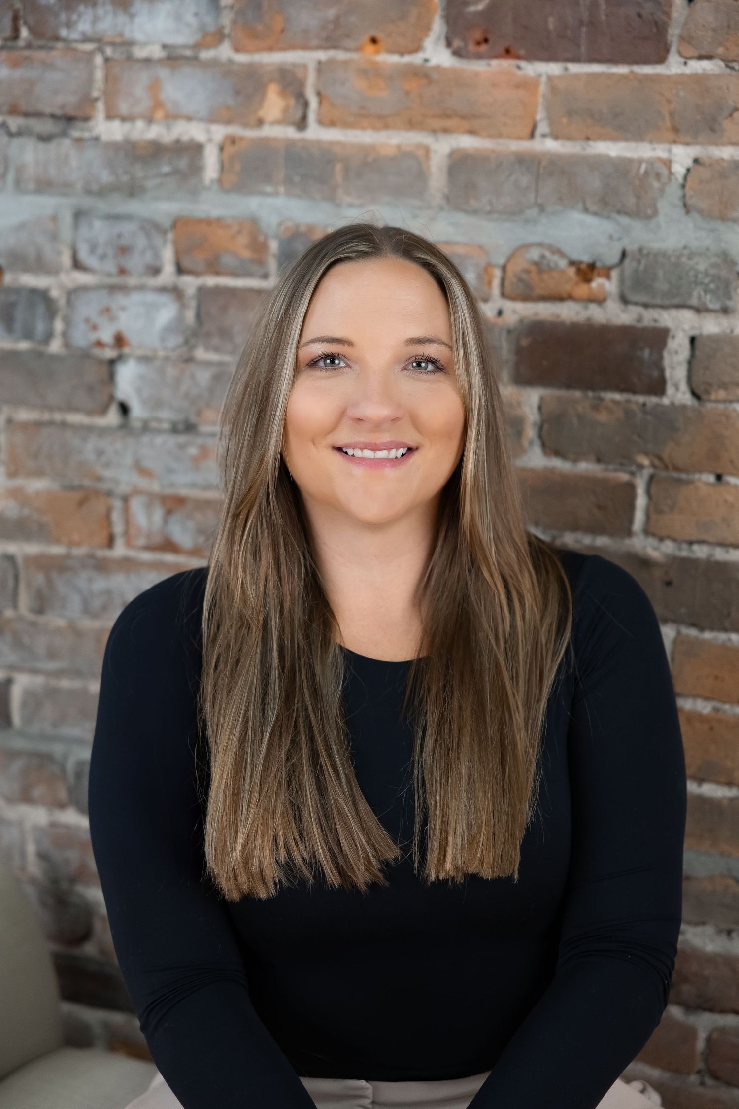 Professional headshot against rustic brick wall showing warm smile and long straight brown hair.