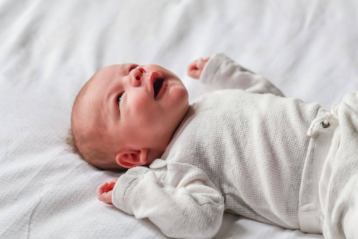 Newborn baby in white clothing sleeping peacefully on white bedding.