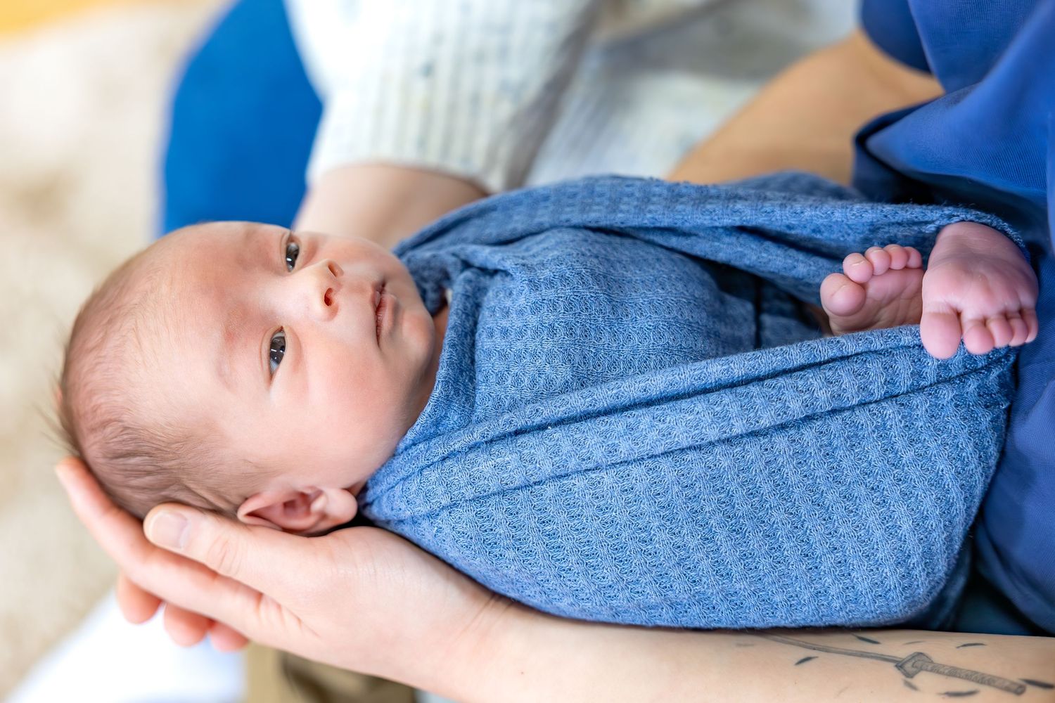 A newborn baby wearing a blue textured onesie lies cradled in gentle hands.