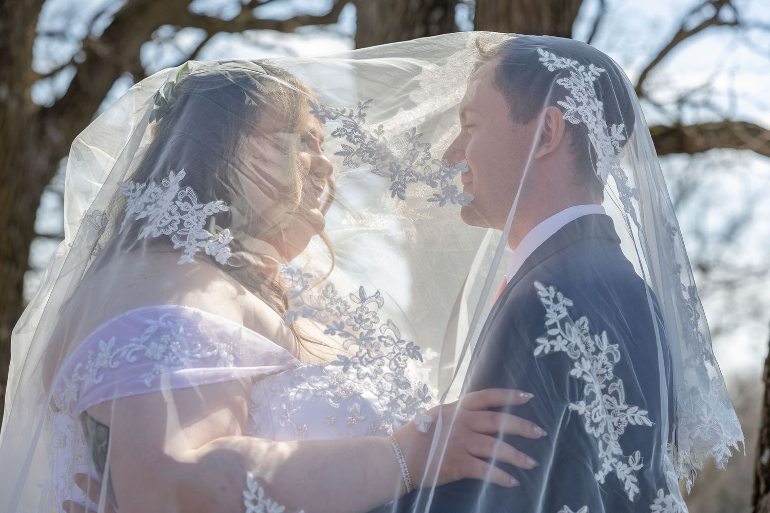 A couple shares a romantic kiss under a flowing white veil during their outdoor wedding ceremony.