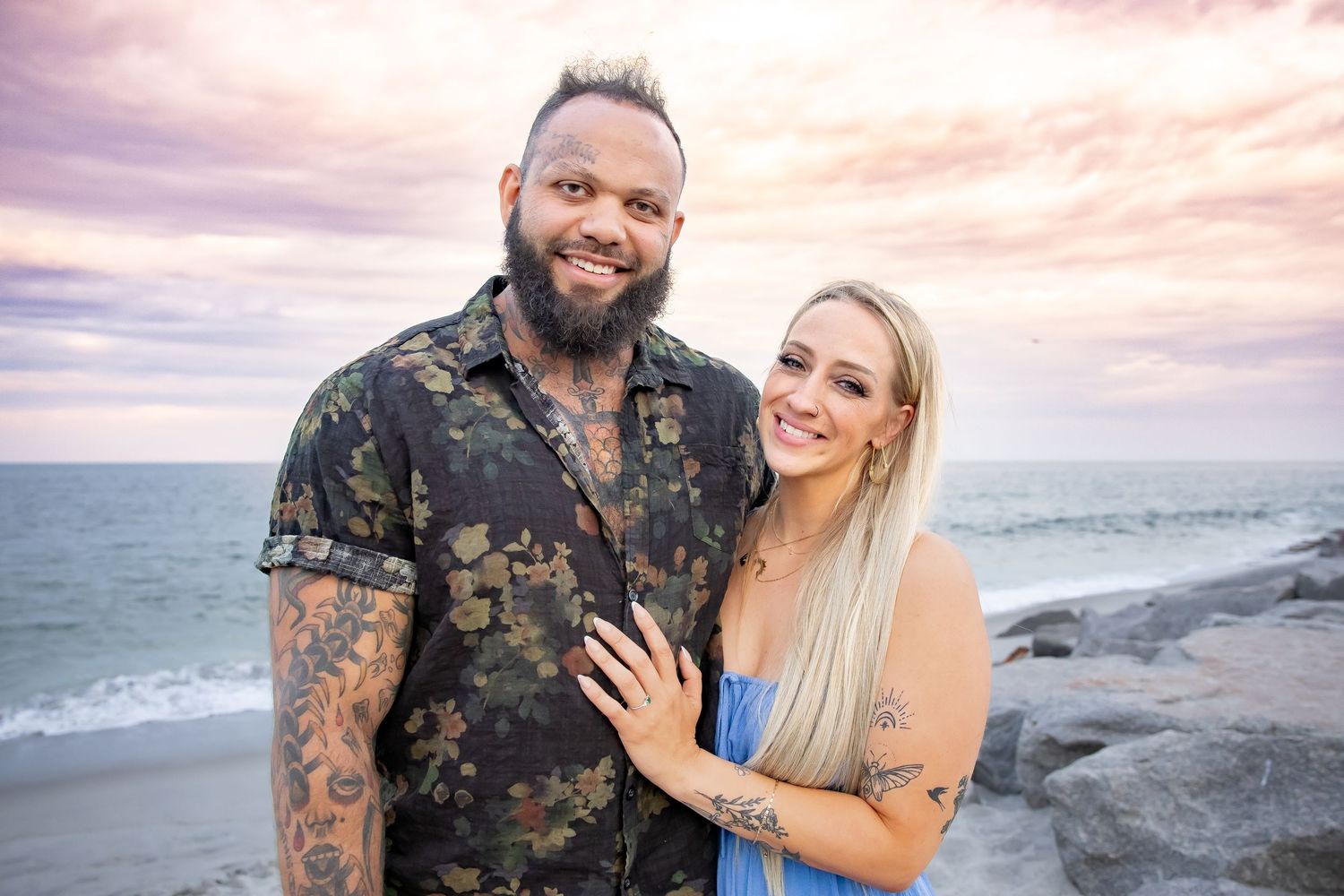 Couple taking engagement photos along rocky coastal beach at sunset.
