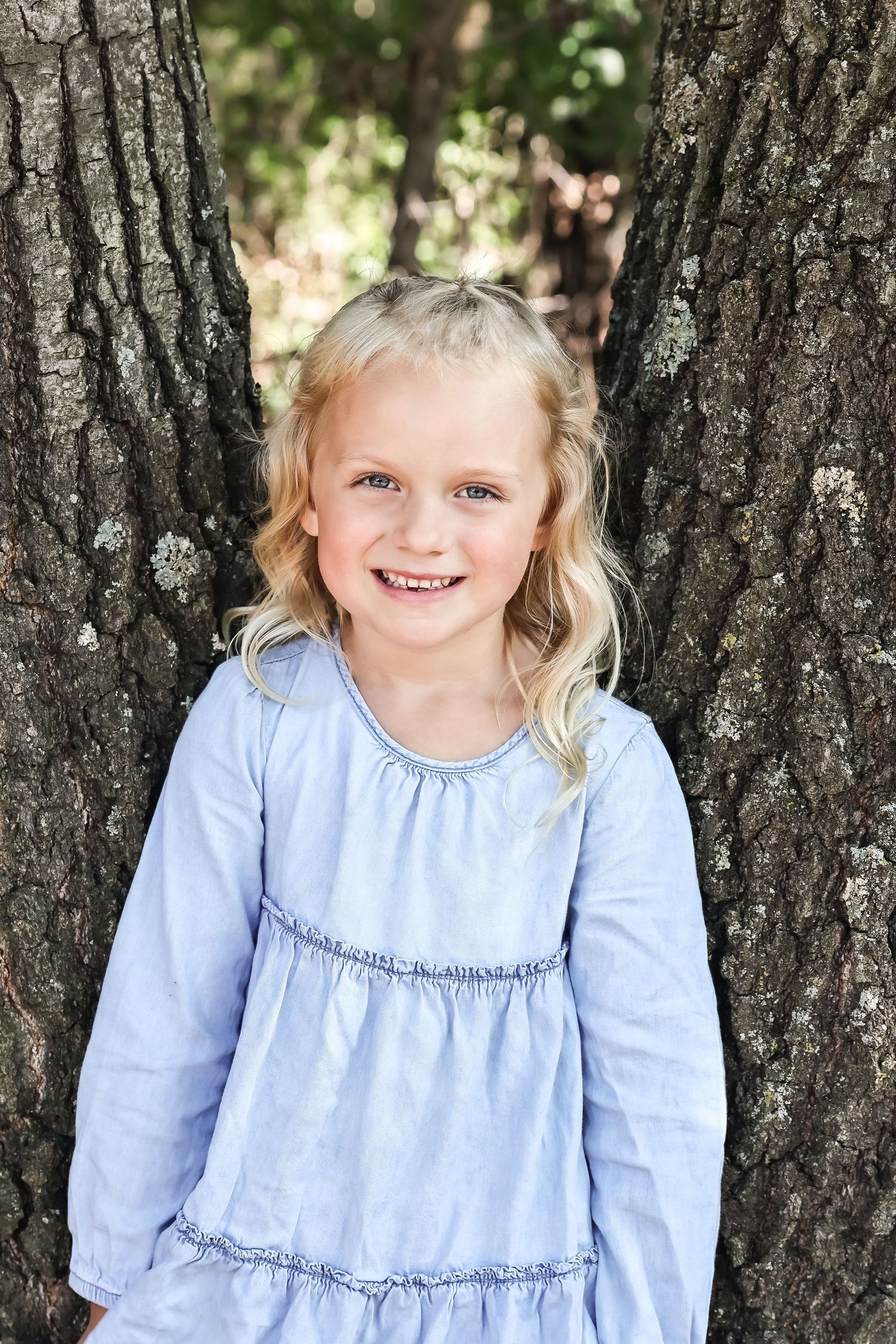 Child in light blue dress poses against textured tree bark background.
