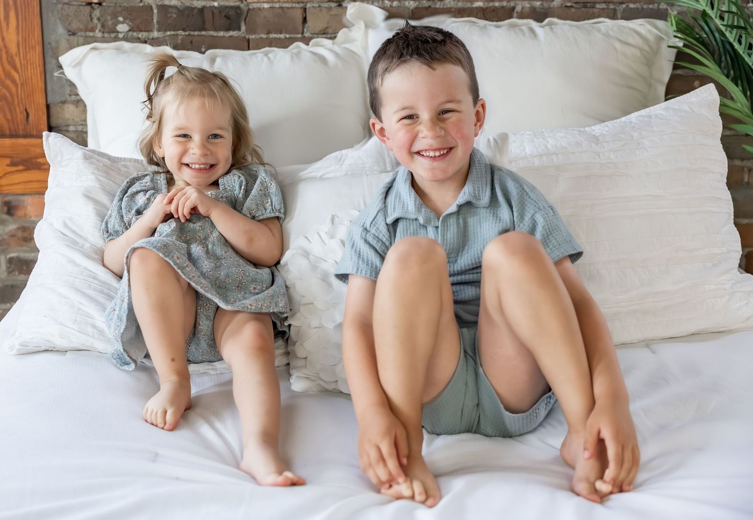 Two siblings in matching blue outfits sit together on white bedding while smiling and showing their bare feet.