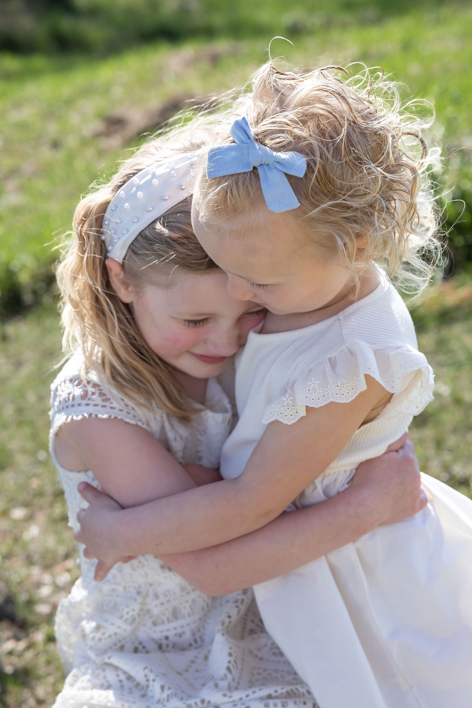Two children in white dresses share a tender embrace outdoors.