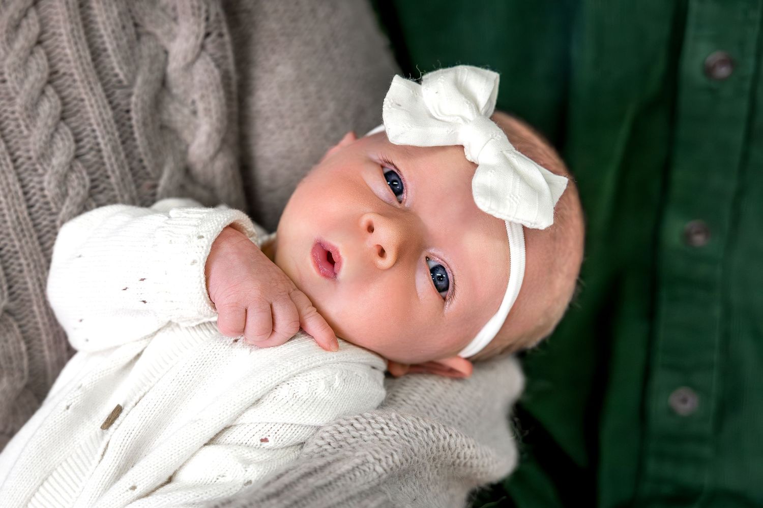 A baby wearing a white bow headband and white outfit lies on a gray blanket.