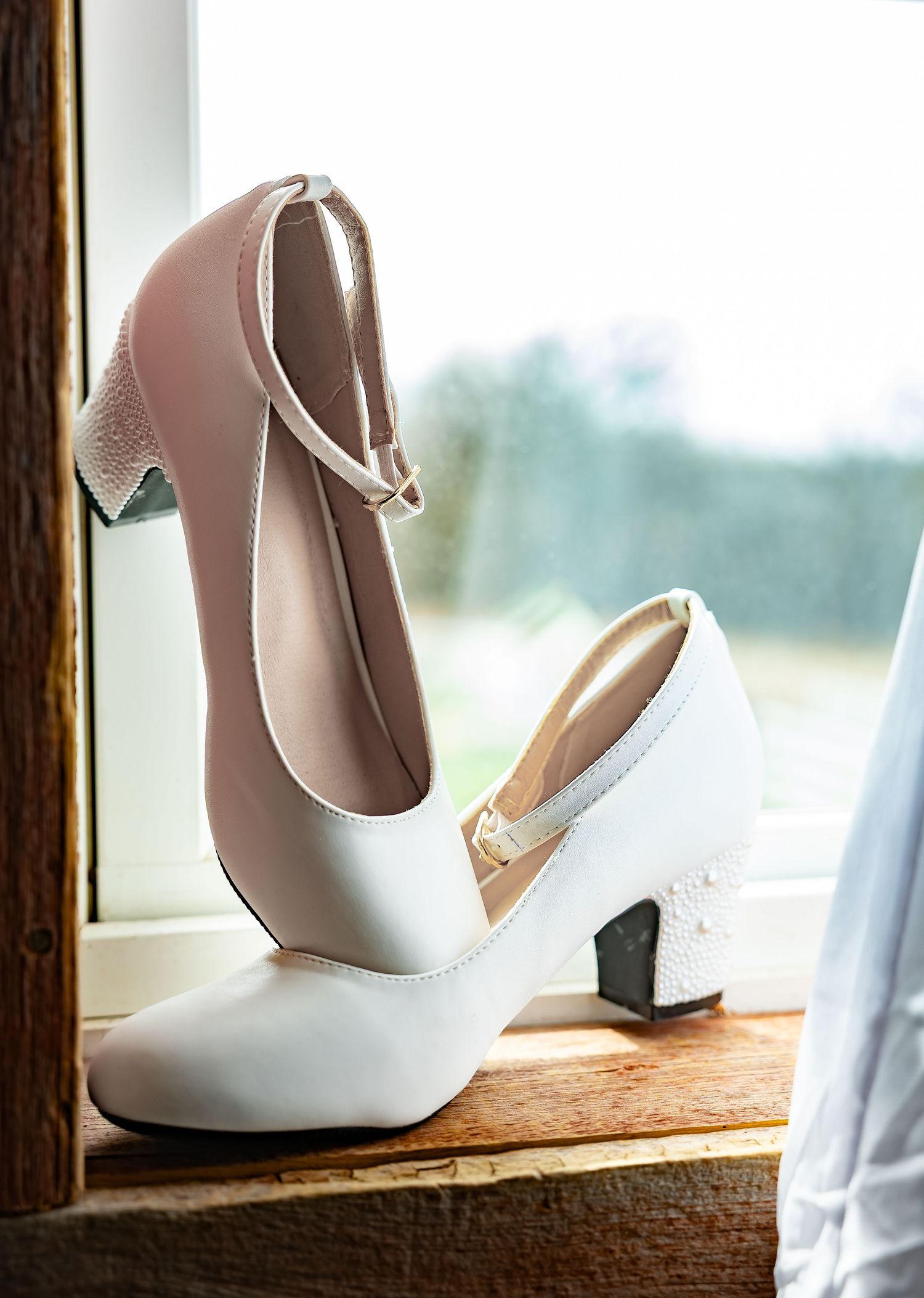 White bridal heels displayed on a wooden surface near a window.