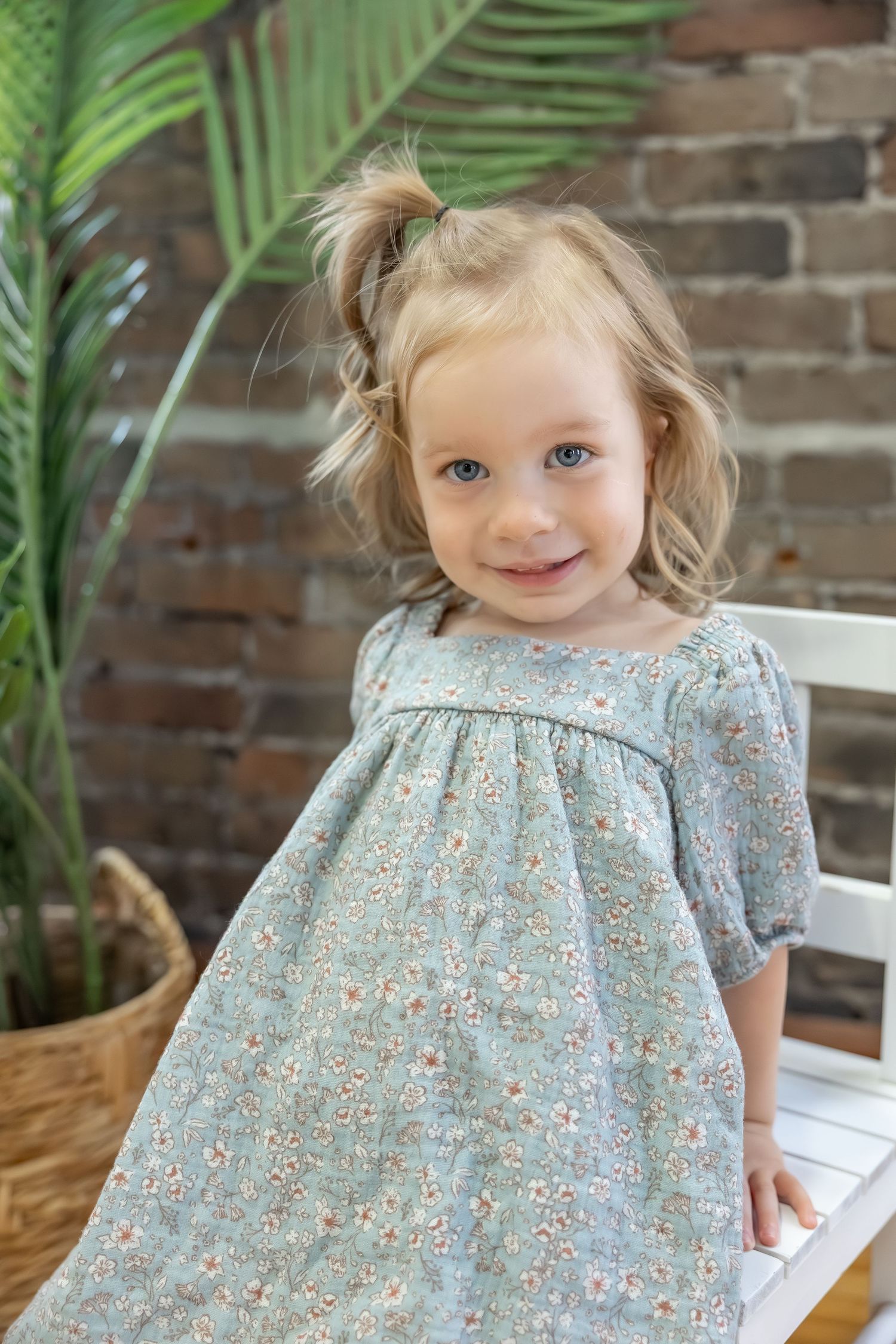 A child in a floral dress poses near green palm leaves and exposed brick wall.
