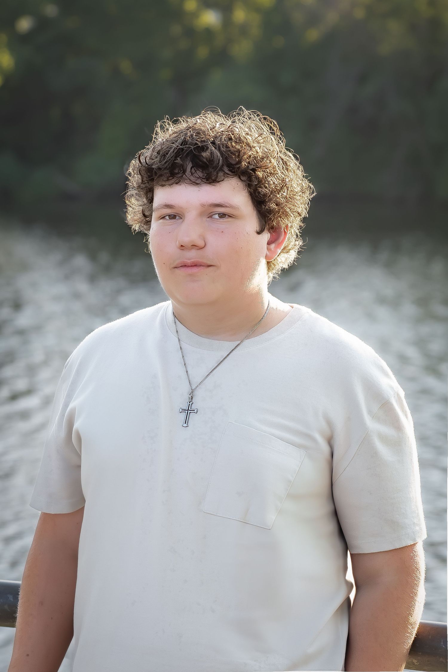 Outdoor portrait with curly hair wearing white t-shirt and cross necklace against misty background.