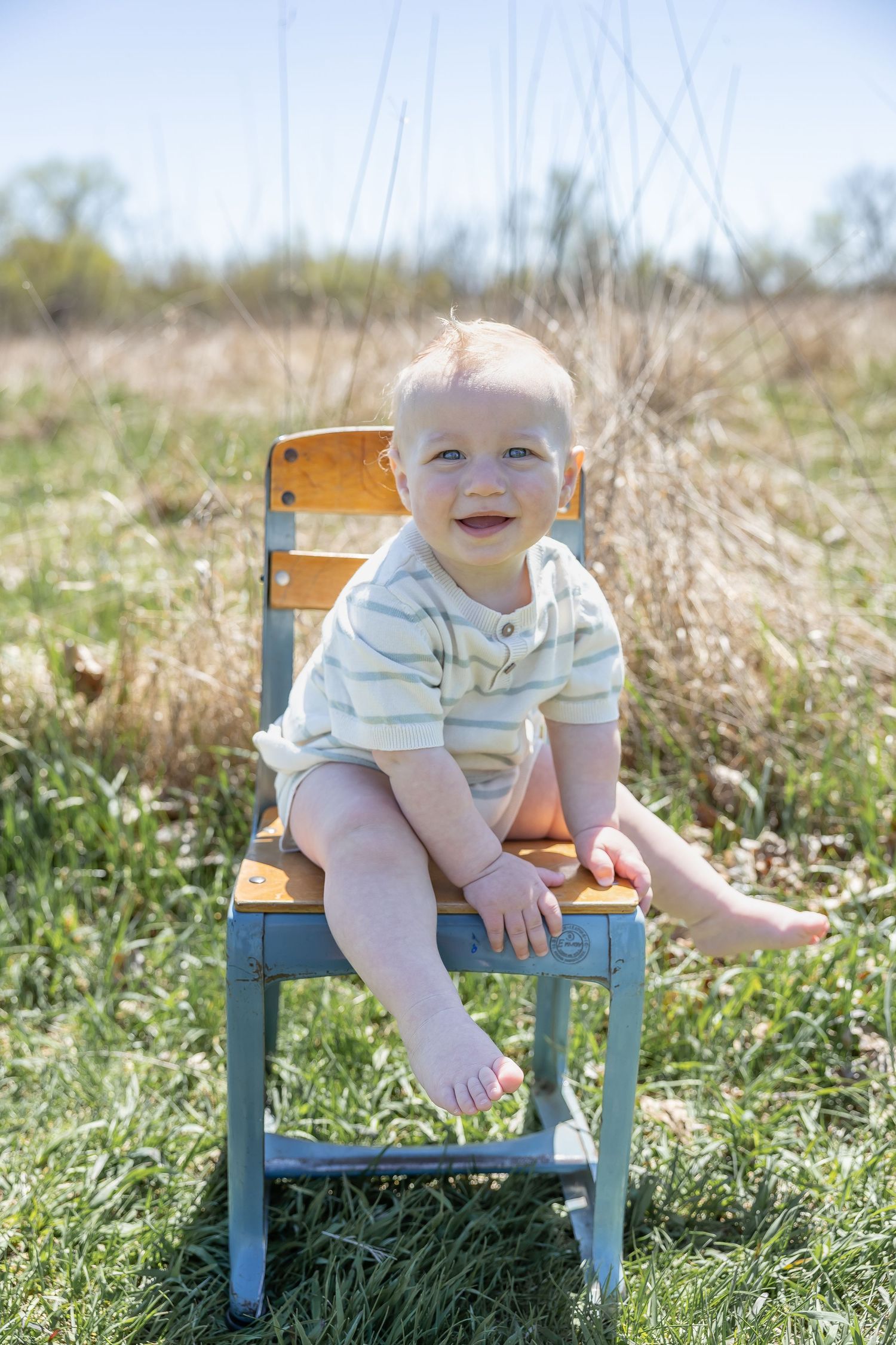 Baby sits on small blue wooden chair in sunny field with tall grass.