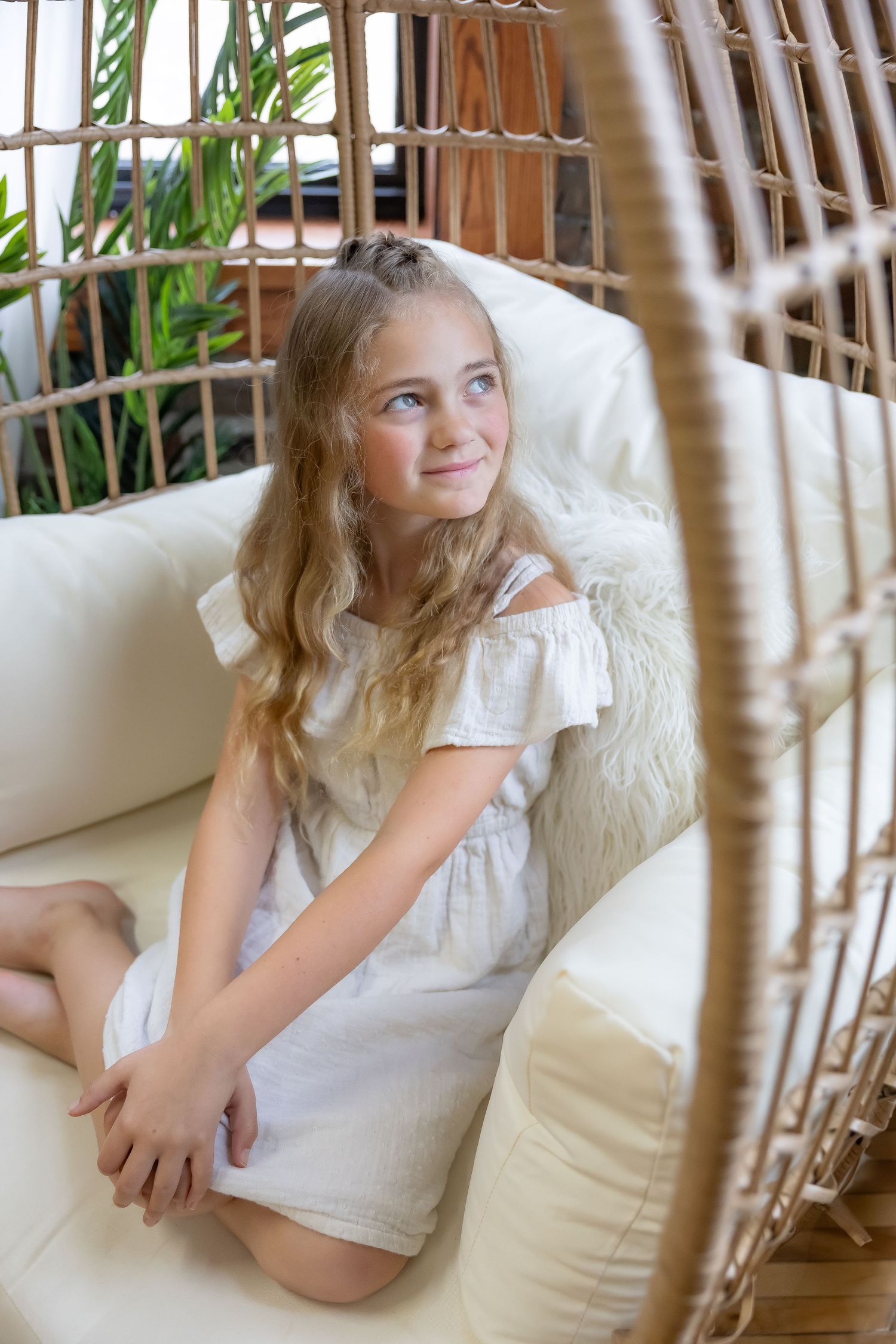Child lounges comfortably in rattan hanging chair wearing white summer outfit.