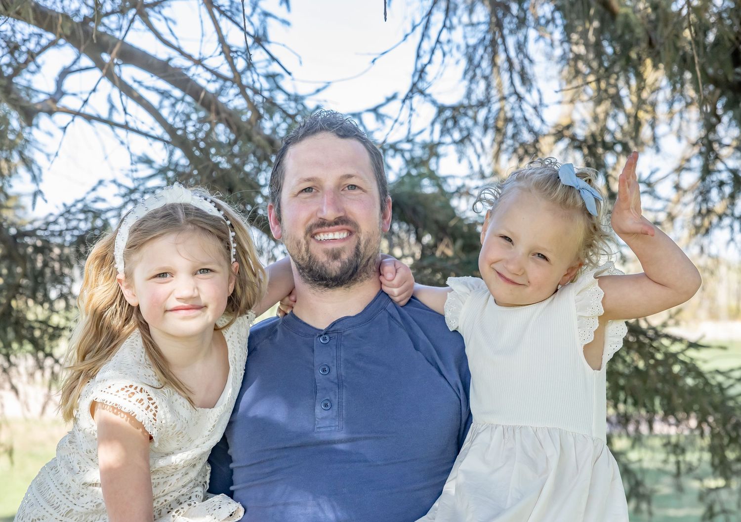 Candid outdoor family portrait with blue sky and evergreen trees in the background.