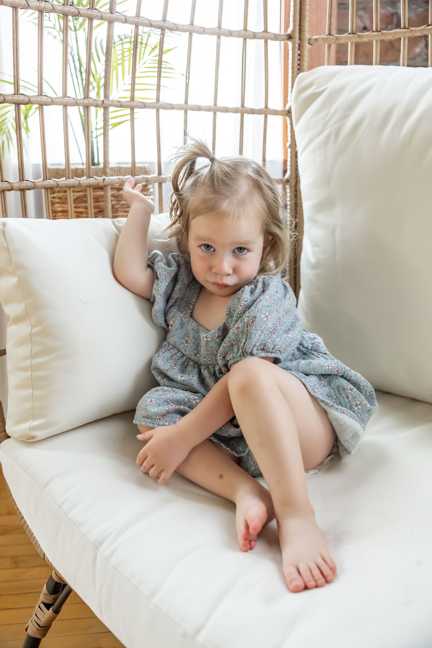 A child in a gray dress relaxes on a white couch near a window with bare feet showing.