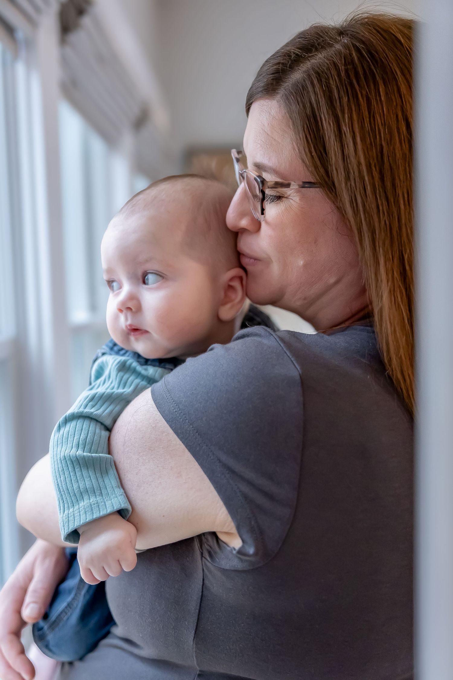 Parent gives gentle kiss to baby while standing by window in intimate portrait.