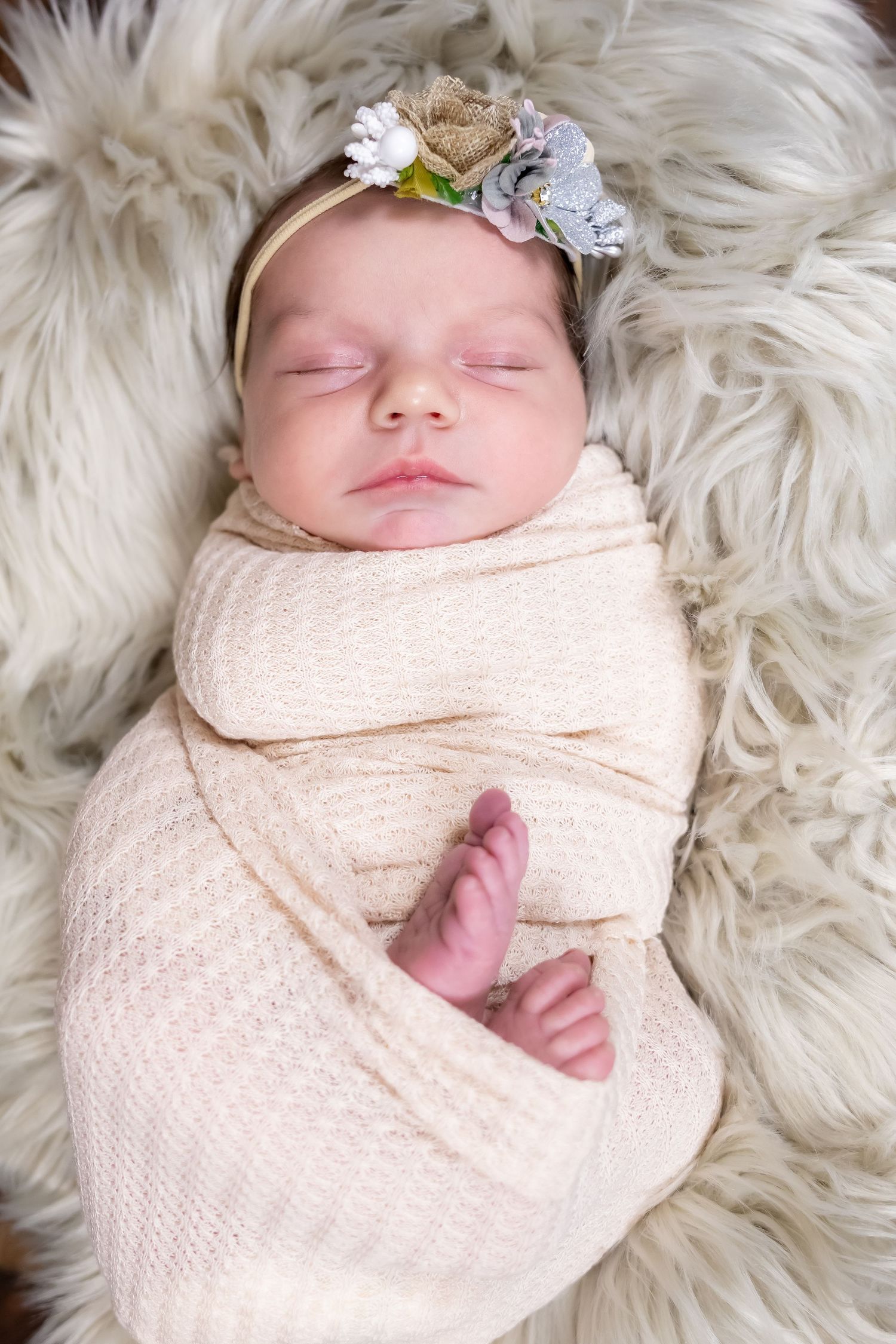 Sleeping newborn wrapped in cream swaddle with floral headband on fluffy white fur backdrop.