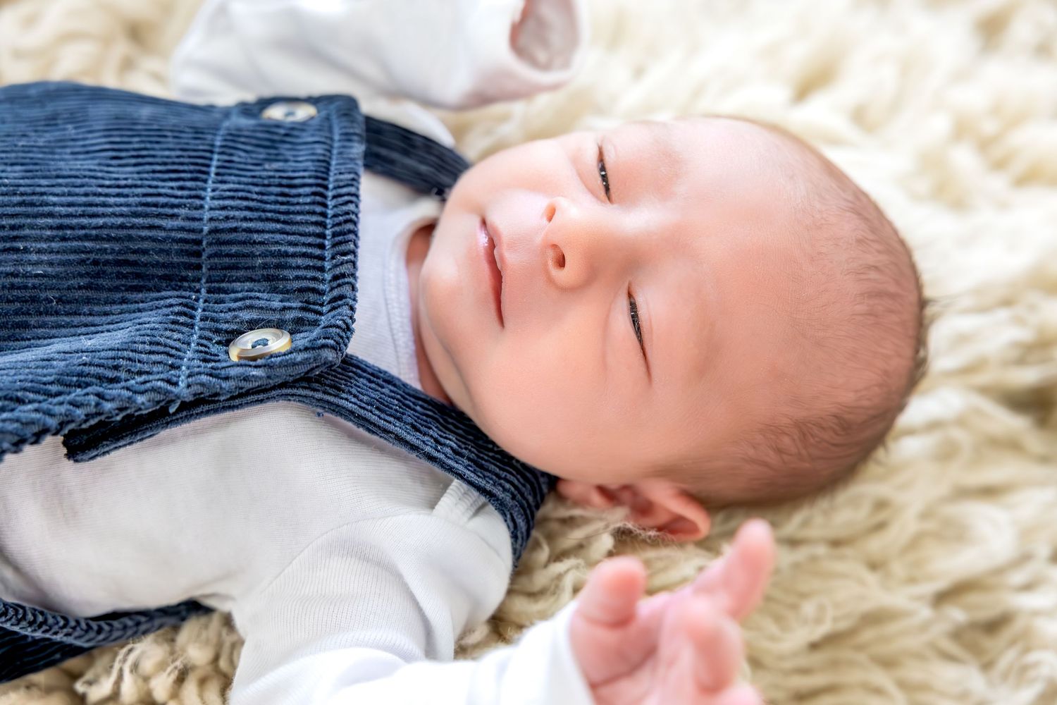 A sleeping baby wearing a white onesie lies on a soft cream-colored fur blanket.